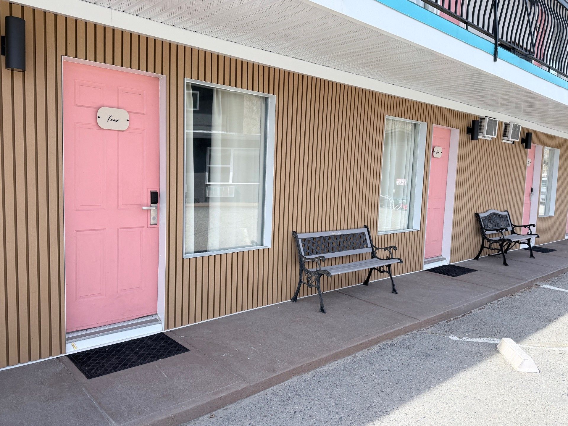 A row of motel units with pink doors, vertical tan wood siding, and metal benches on a concrete walkway.