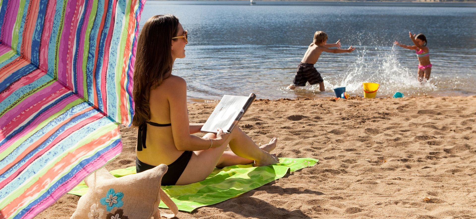 A person sits on a lime-green beach towel reading a book under a striped umbrella while two children play in the water.