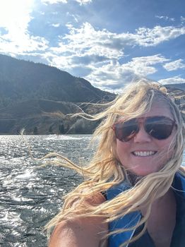A smiling person with long, windblown blonde hair and sunglasses on a boat, with mountains and a sparkling lake behind.
