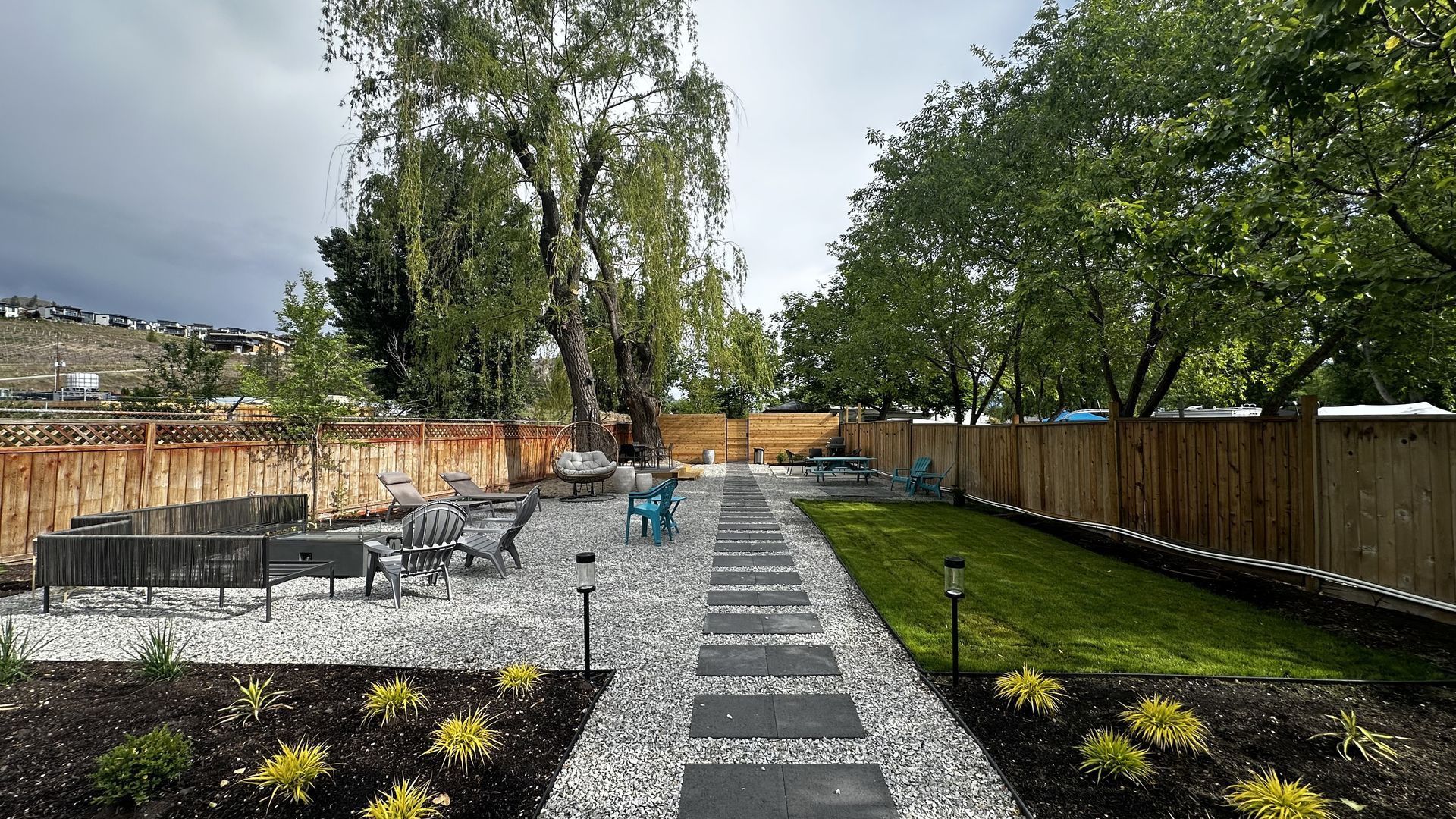 A backyard with a stone paver path, white gravel patio area with outdoor chairs, a small green lawn, and wood fencing.