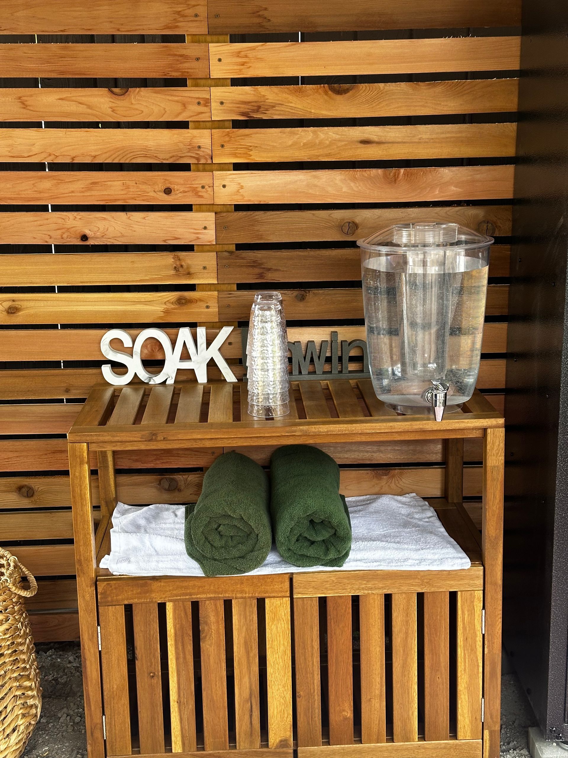 A wooden shelving unit against a wood-slatted wall holding a water dispenser, stack of cups, and two dark green towels.