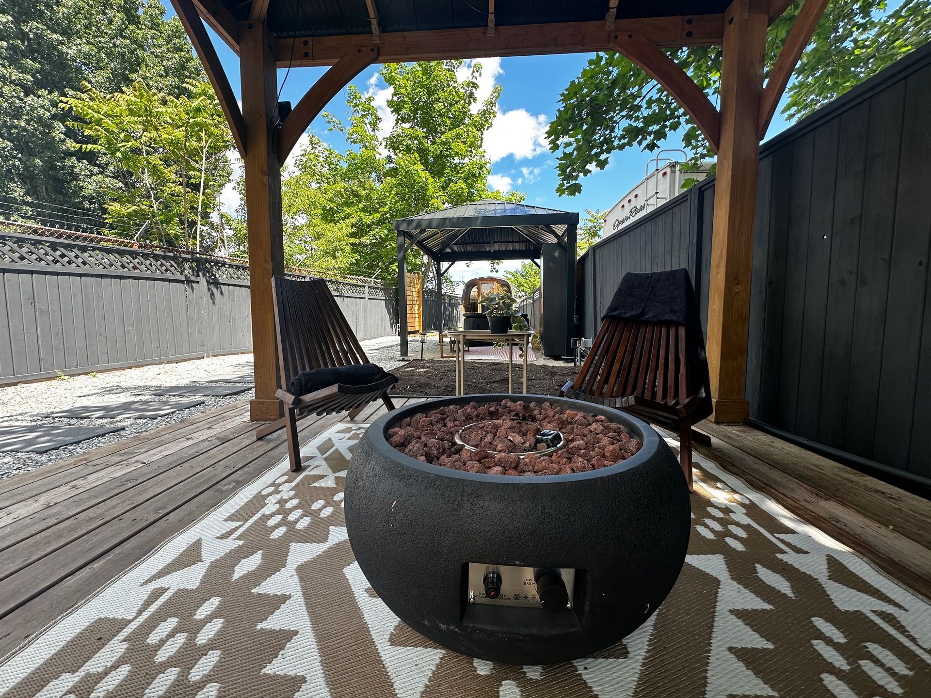 A round fire pit sits on a patterned rug under a wooden pergola, with two chairs and a fenced backyard in the background.