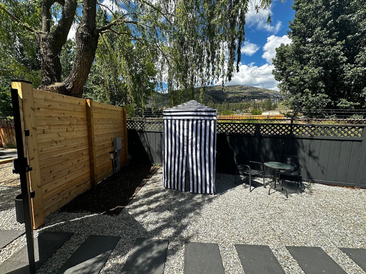 A gravel patio featuring a privacy fence, a blue and white striped outdoor shower enclosure, and a small bistro set.