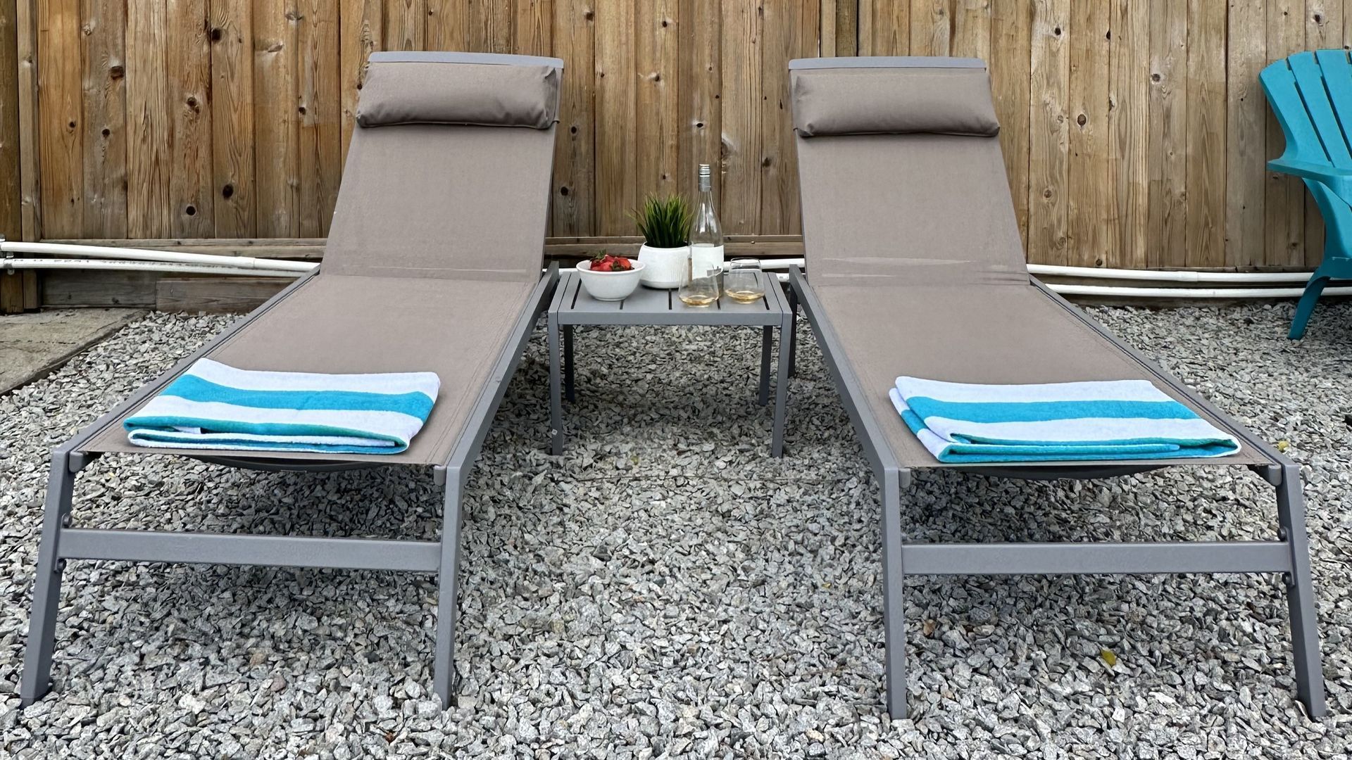Two taupe lounge chairs with folded blue and white striped towels, separated by a small table with a plant, on gravel.