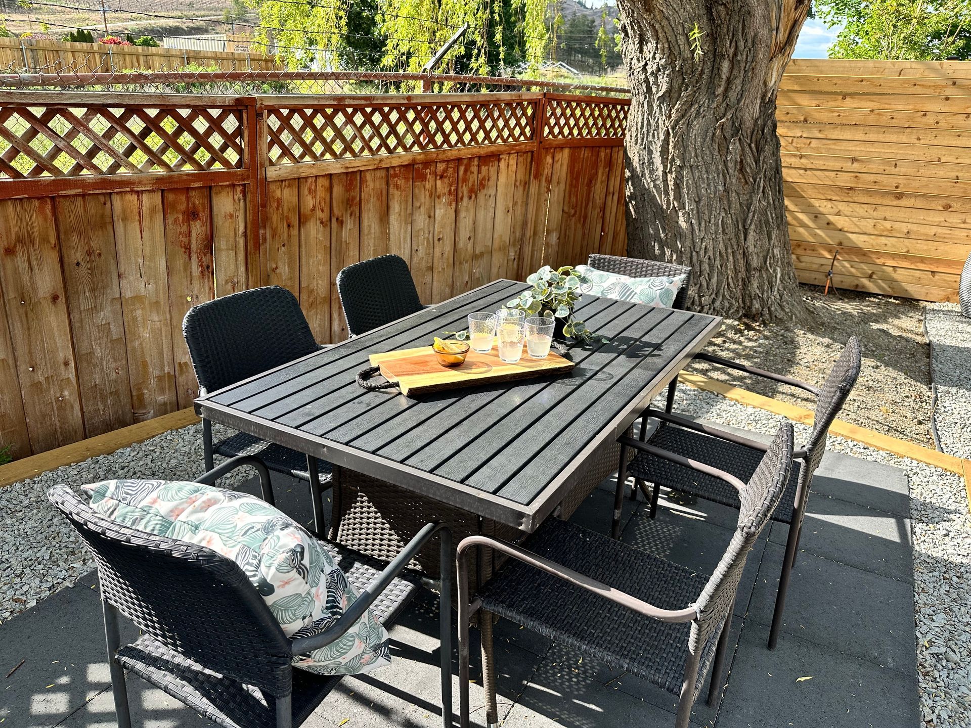 A black patio dining set with six chairs on a gravel area next to a wooden fence and a large tree.