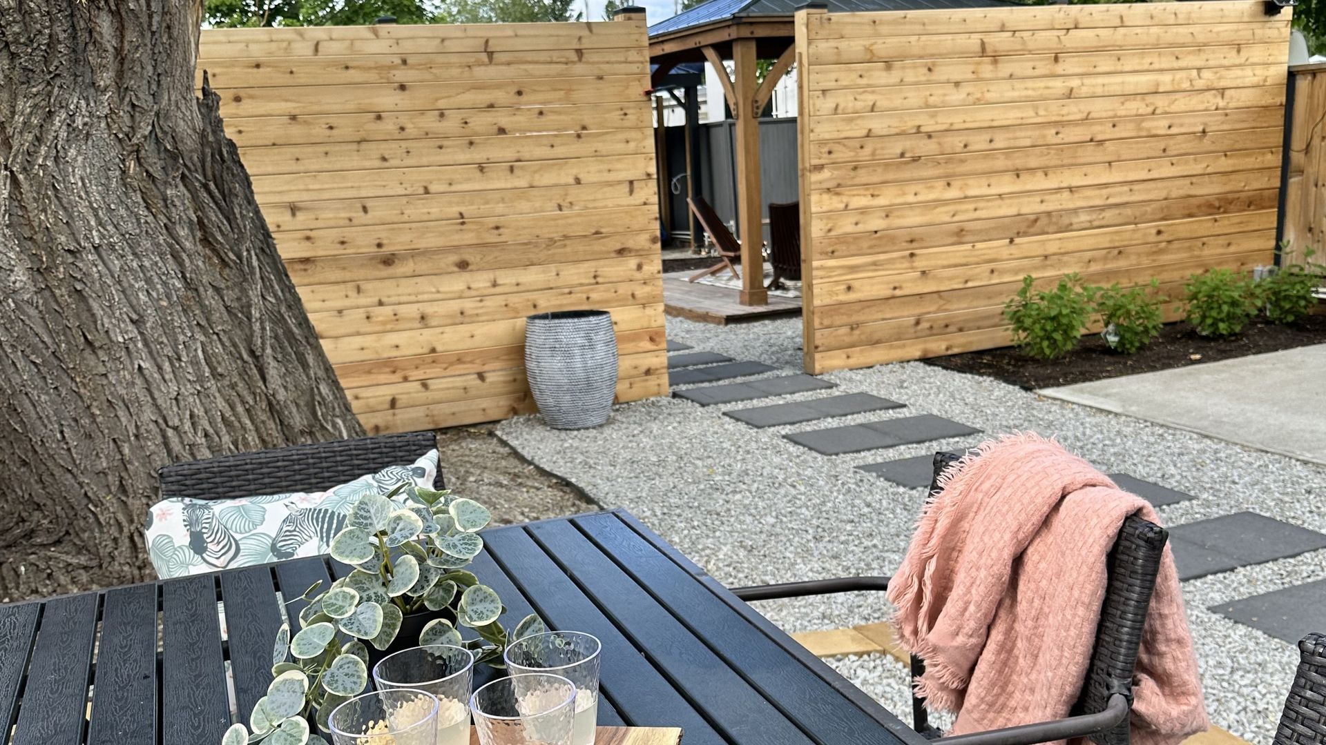 A wooden fence opens to a gravel path with stone pavers, leading to a backyard gazebo under a large tree.