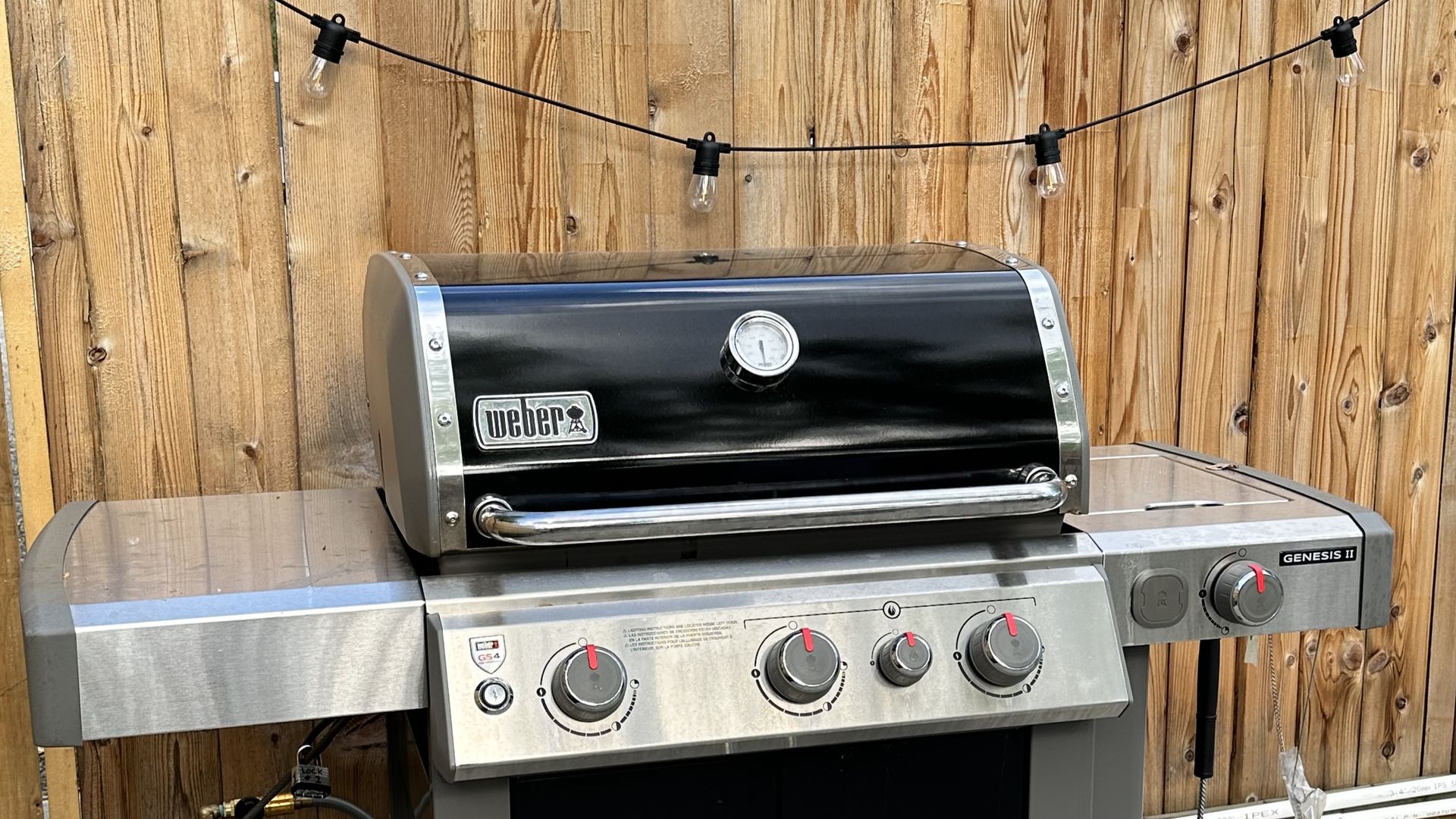 A black and stainless steel Weber gas grill sits outdoors against a wooden fence with decorative string lights.