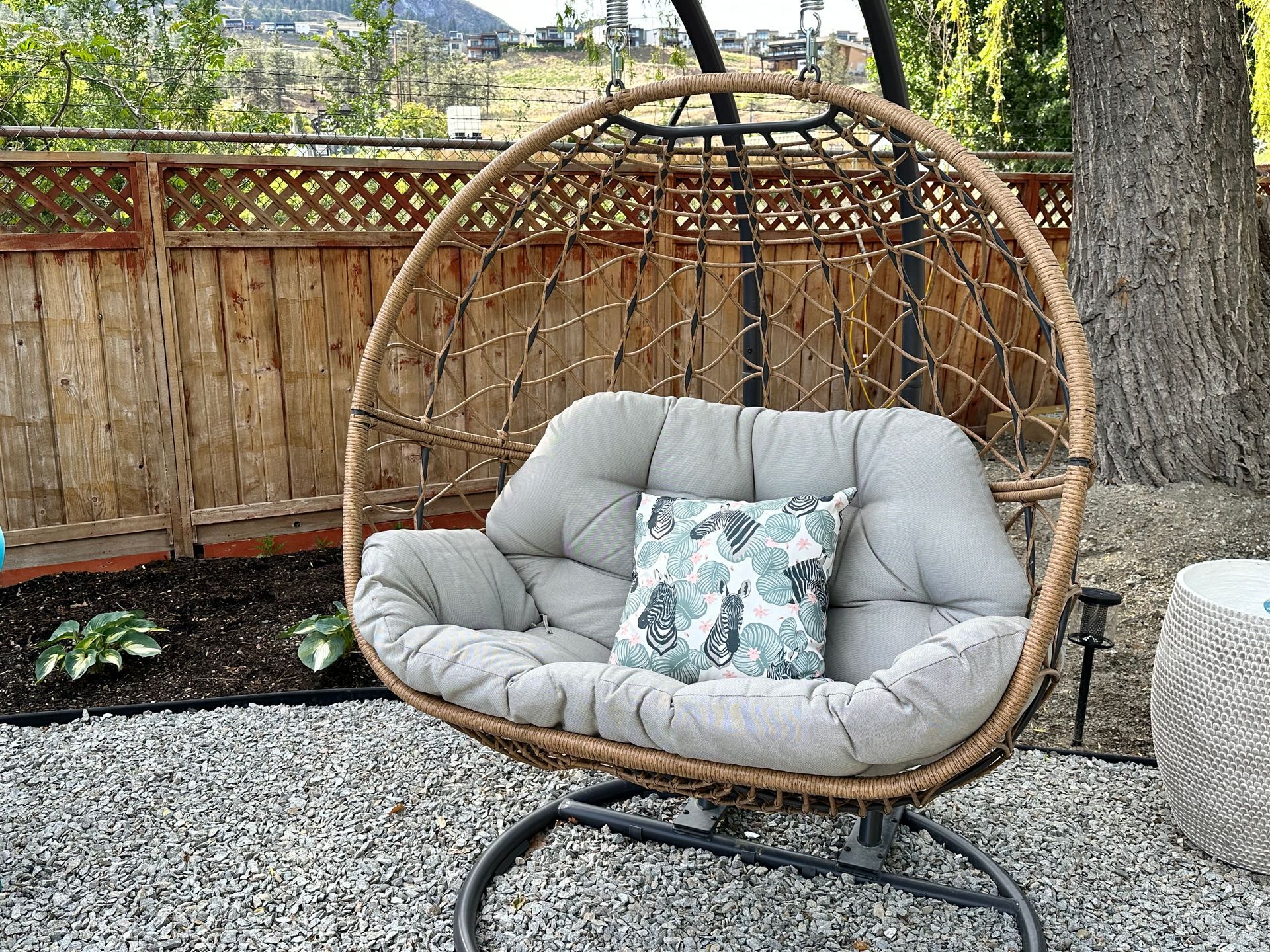 A beige wicker hanging egg chair with a gray cushion and a patterned throw pillow, set on a gravel patio by a fence.