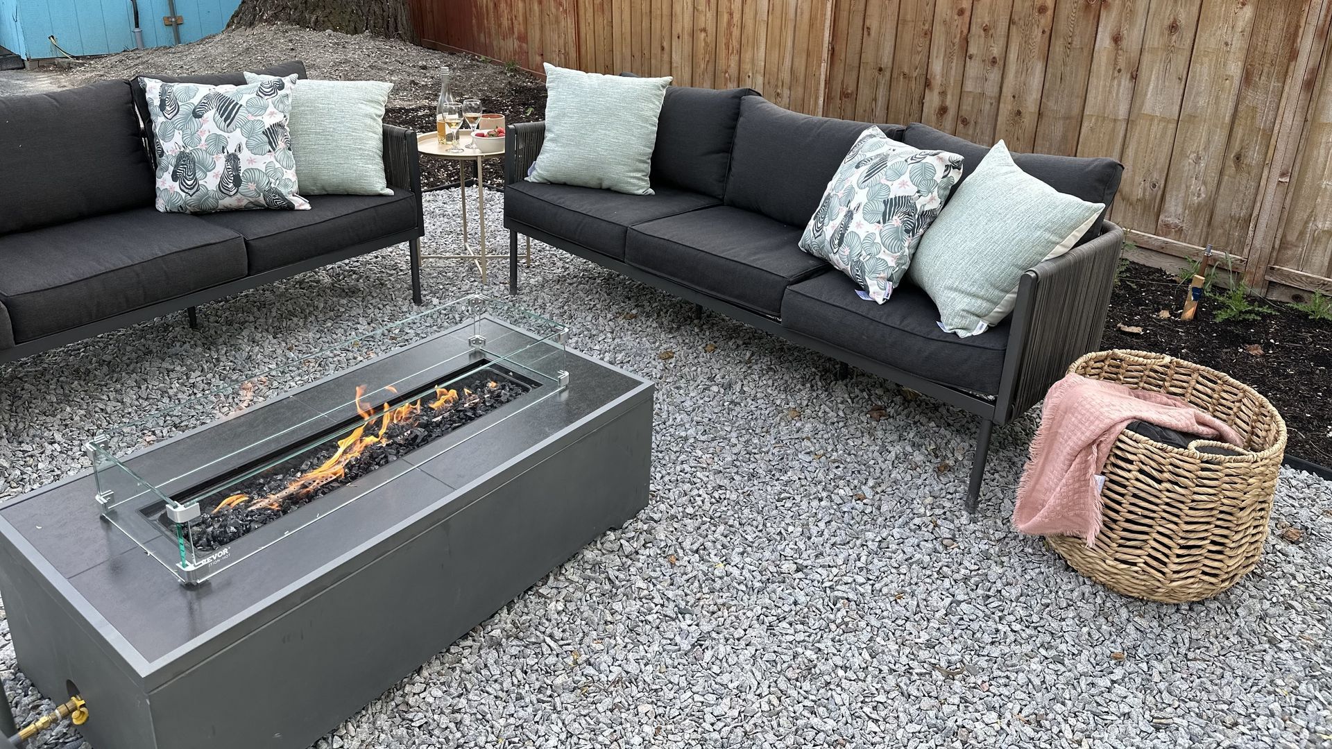 Outdoor patio featuring charcoal sofas with decorative pillows, a lit rectangular fire pit, and a woven basket on gravel.