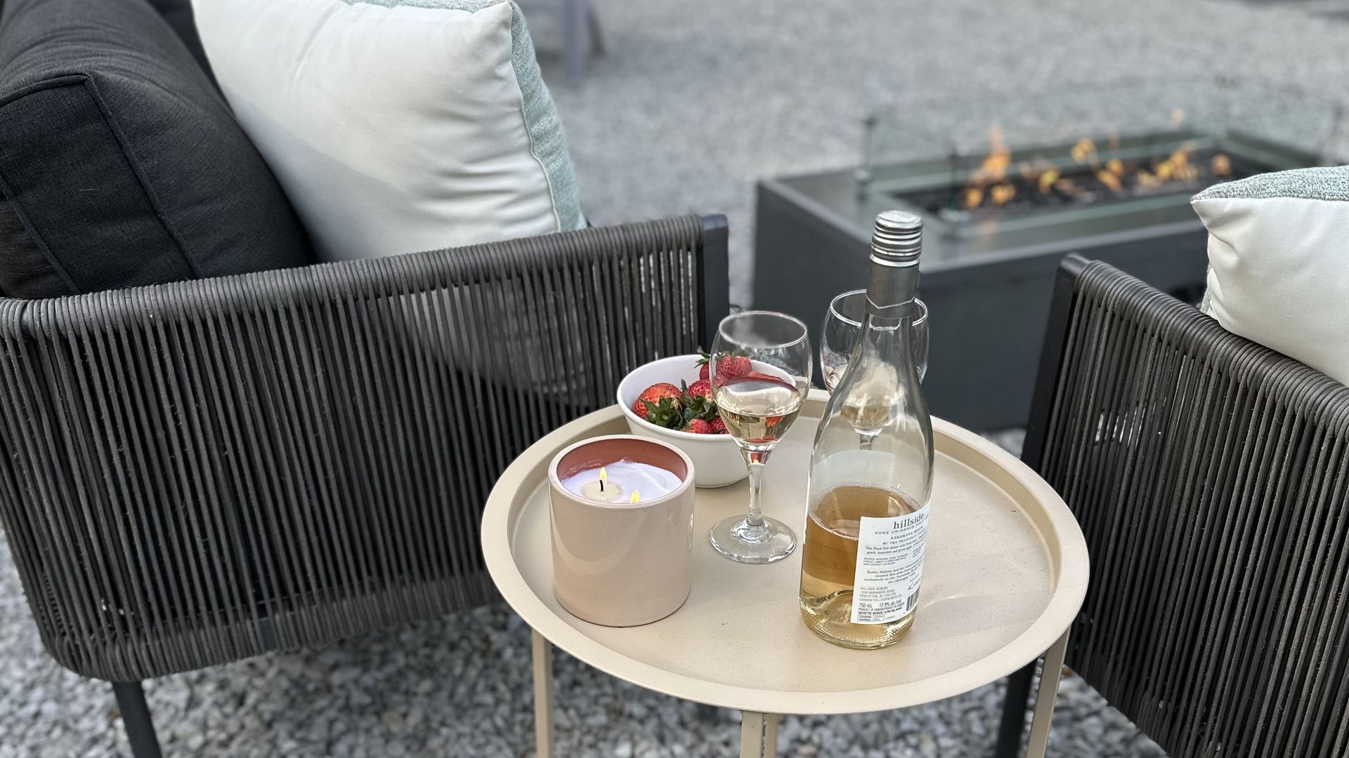 A patio table holds a candle, strawberries, and a bottle of wine with a glass near a lit outdoor fire pit.