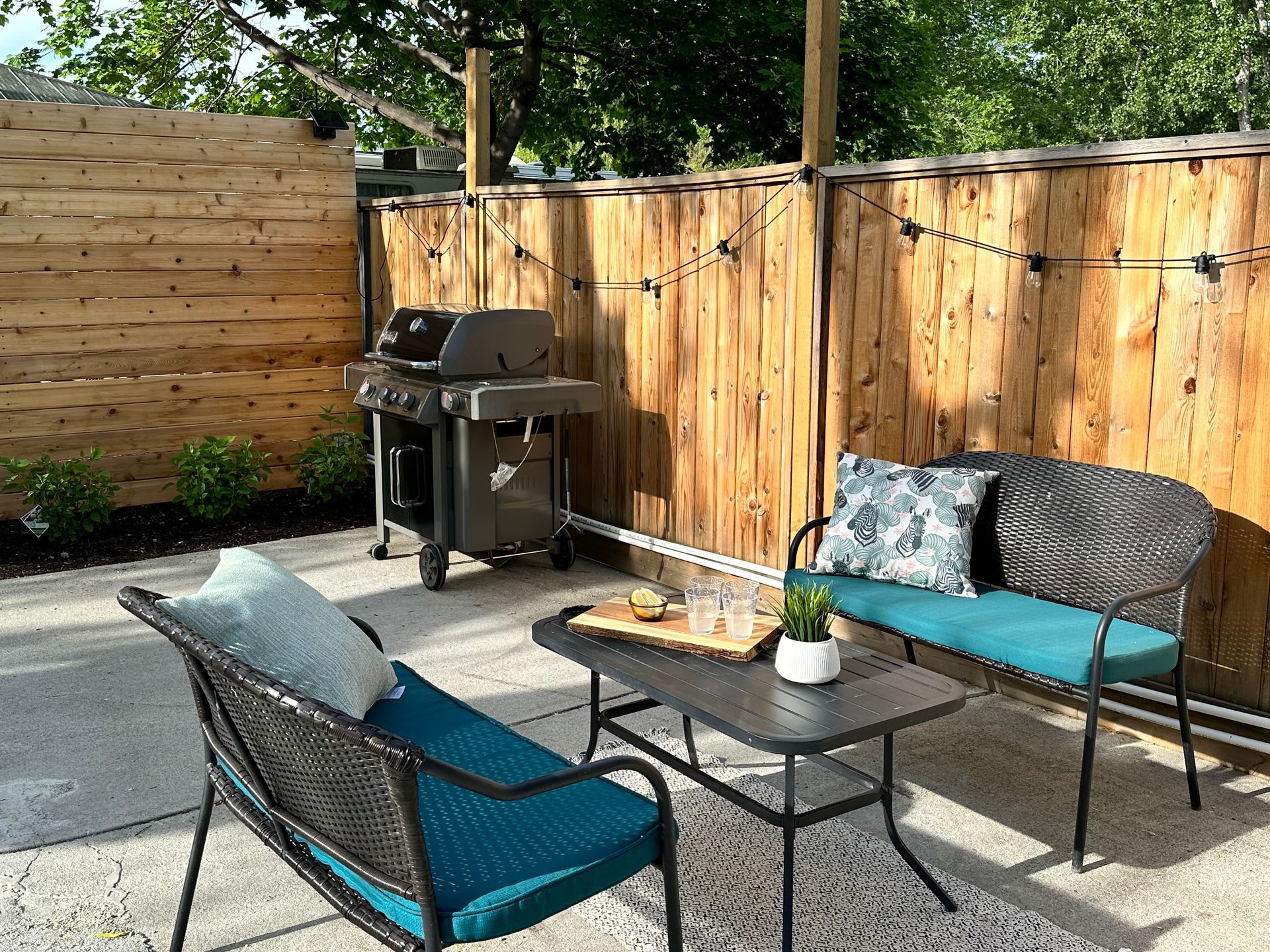 A cozy patio with two chairs, a small table, a grill, and wooden privacy fencing decorated with string lights.