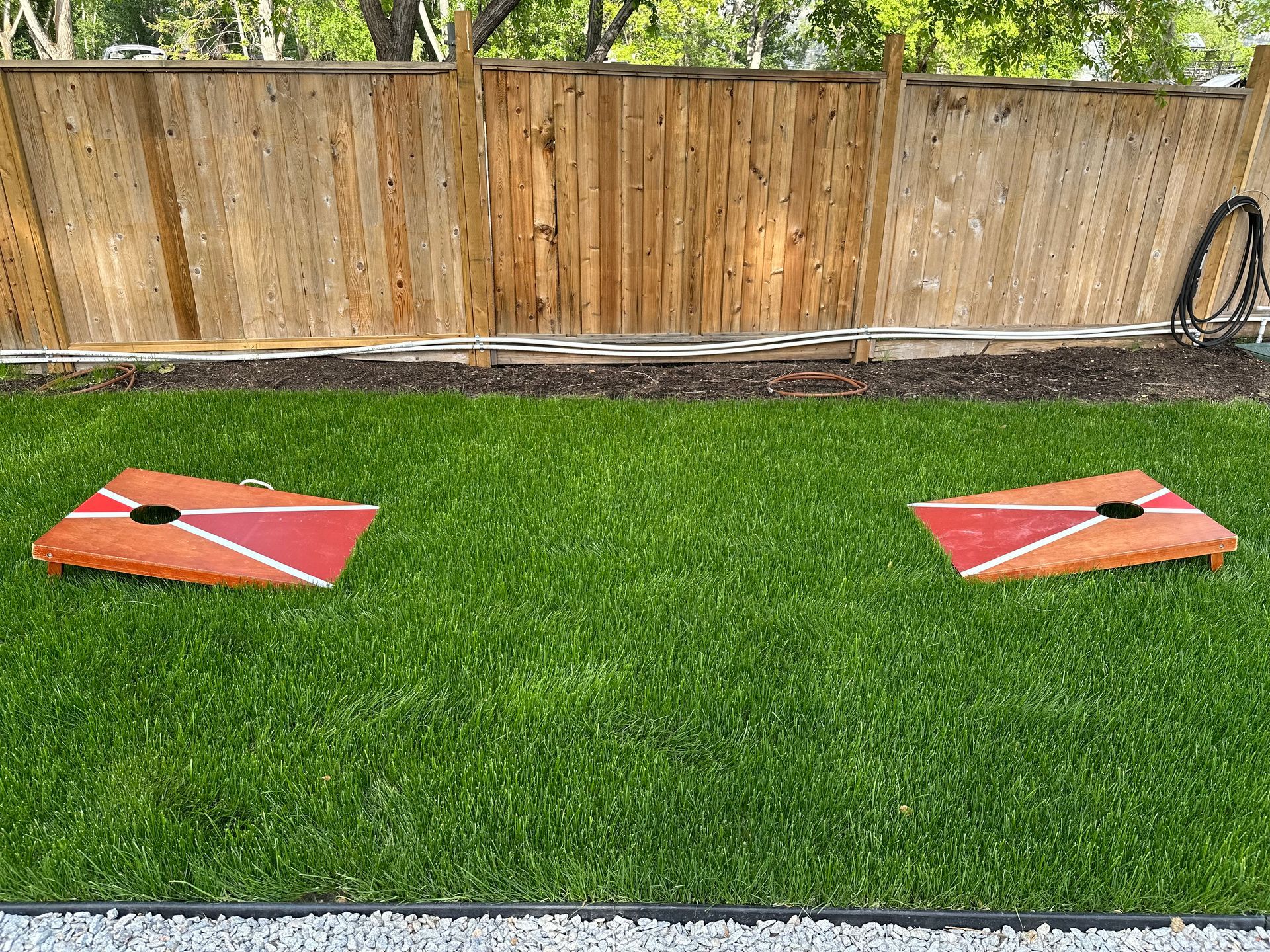 Two red and white painted cornhole boards set up on a grassy lawn in front of a wooden fence.