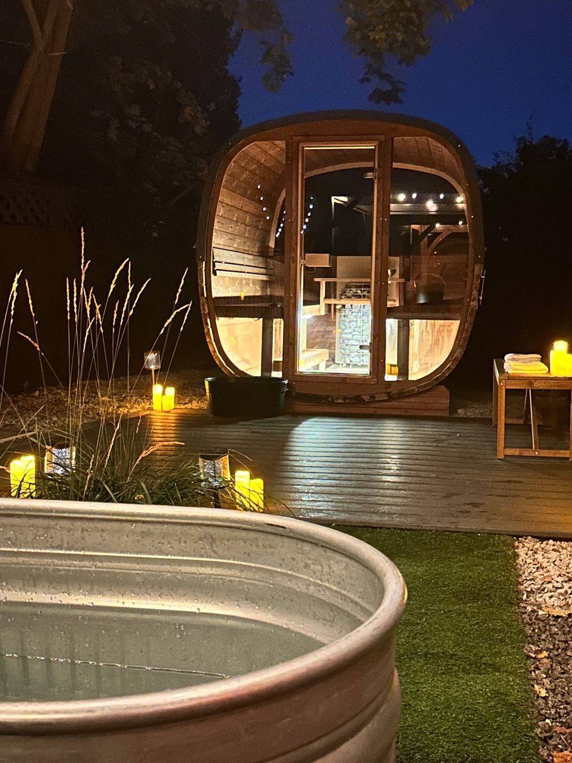 A barrel-shaped wooden sauna illuminated at night next to a galvanized metal stock tank pool on a patio with candles.