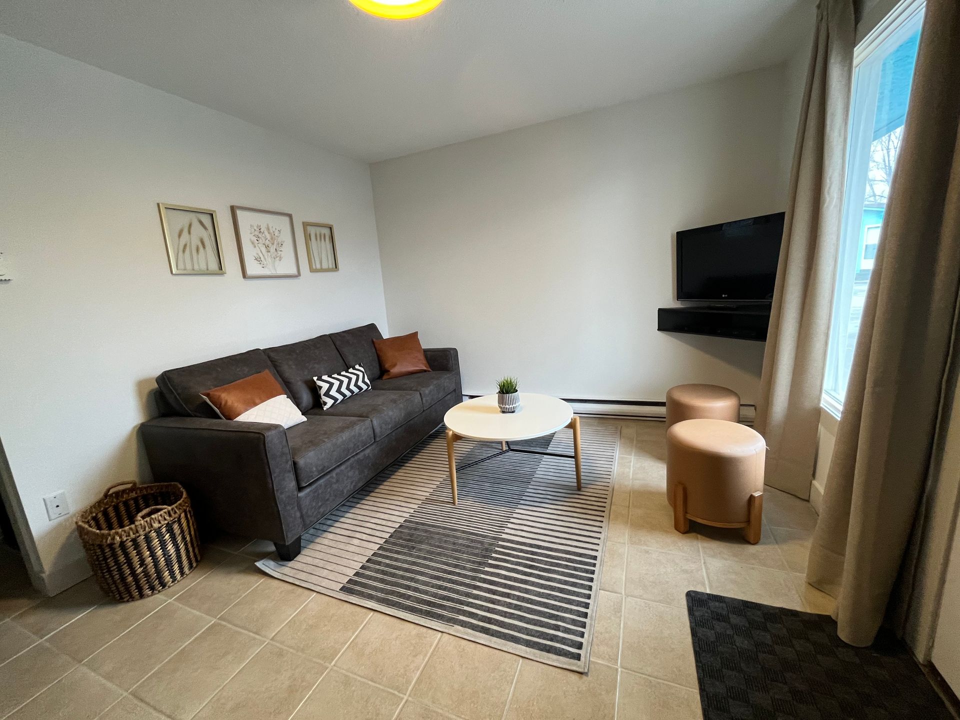 A modern living room with a dark grey sofa, patterned area rug, round coffee table, two tan ottomans, and a wall-mounted TV.