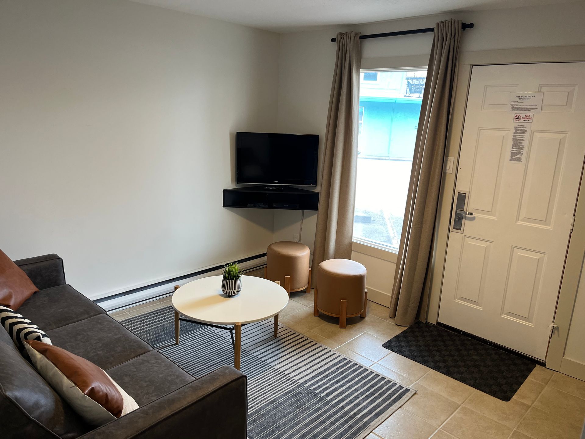 A cozy living room with a dark sofa, a round coffee table on a patterned rug, a wall-mounted TV, and a white entrance door.