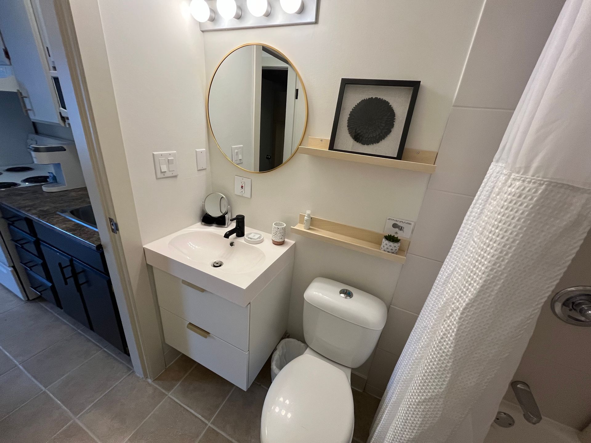 A small, modern bathroom with a white pedestal sink, round gold-rimmed mirror, toilet, and light-wood wall shelves.