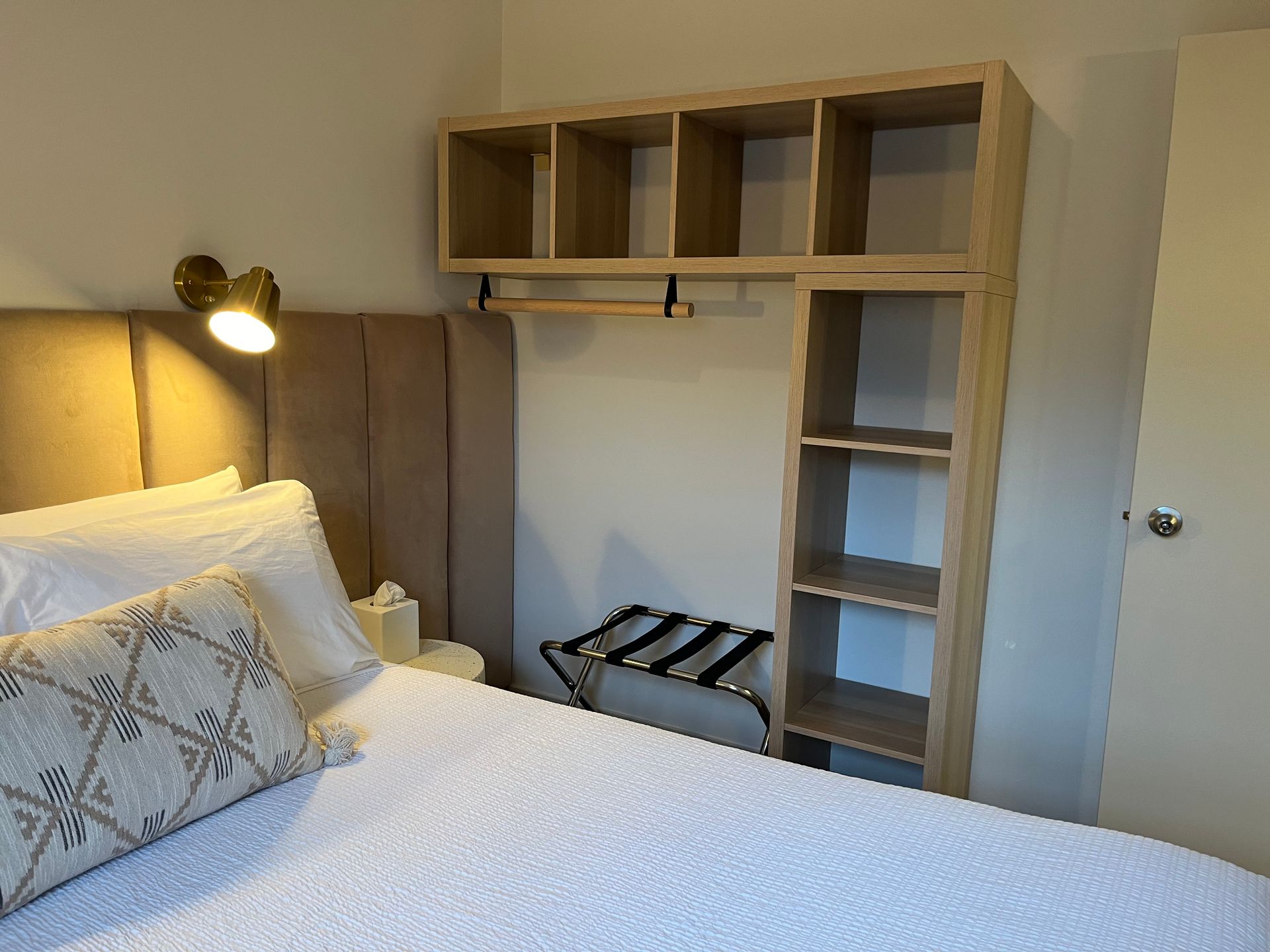 A hotel bedroom featuring a bed with a tan upholstered headboard, a wall-mounted light, and wooden open shelving.
