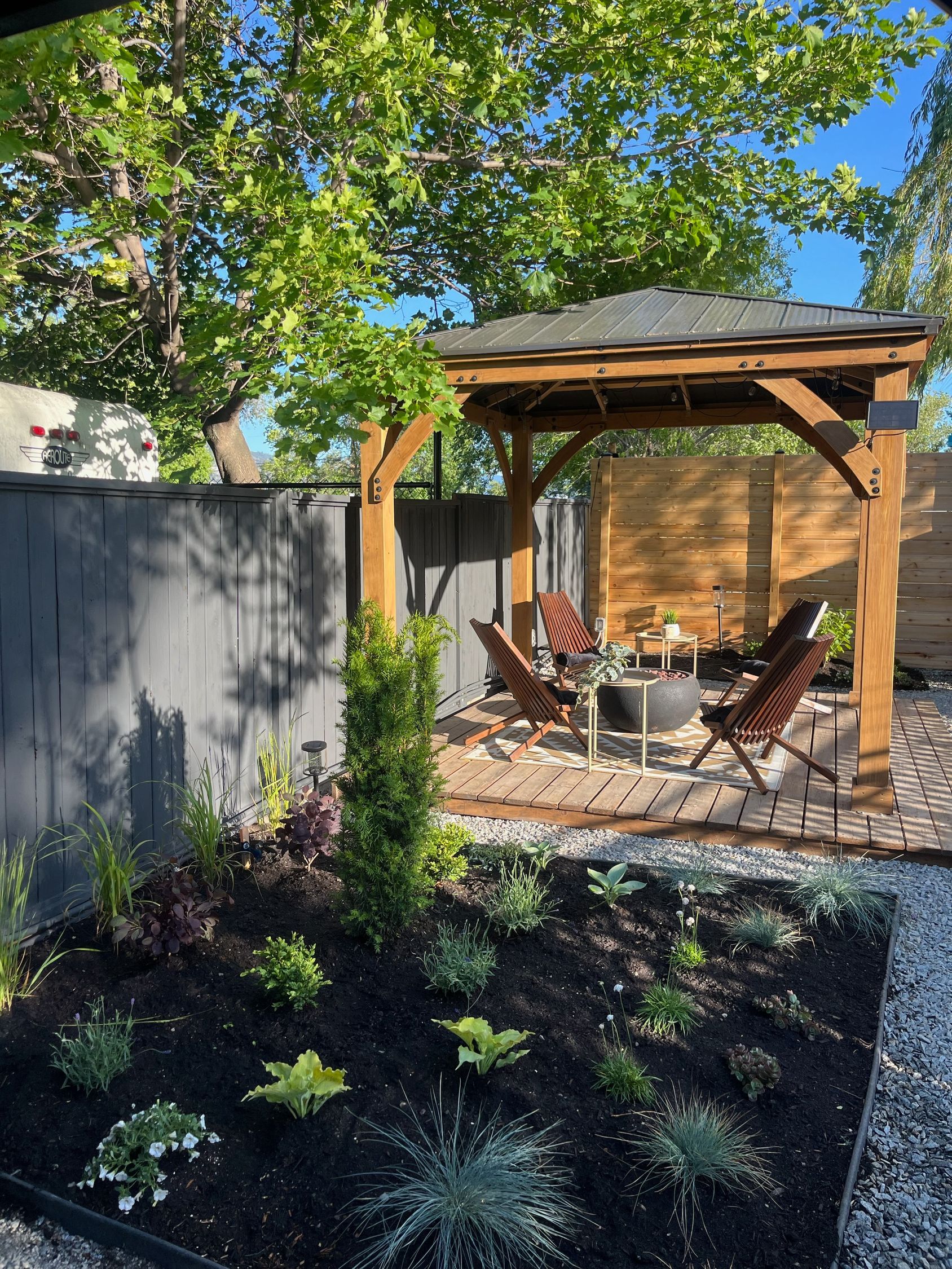 A wooden pergola with patio chairs and a fire pit sits on a stone base, next to a landscaped garden bed with dark mulch.