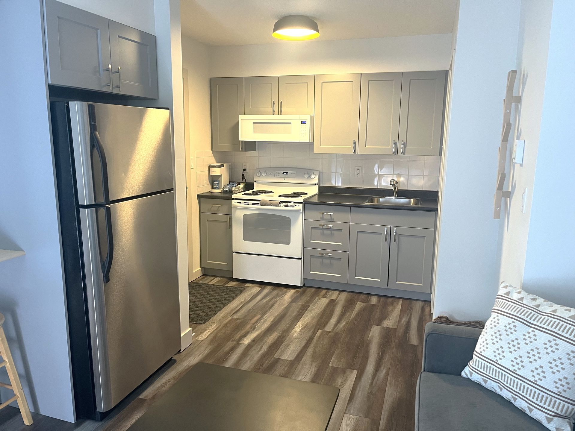 A compact kitchen with grey cabinets, stainless steel refrigerator, white stove, and wood-patterned flooring.