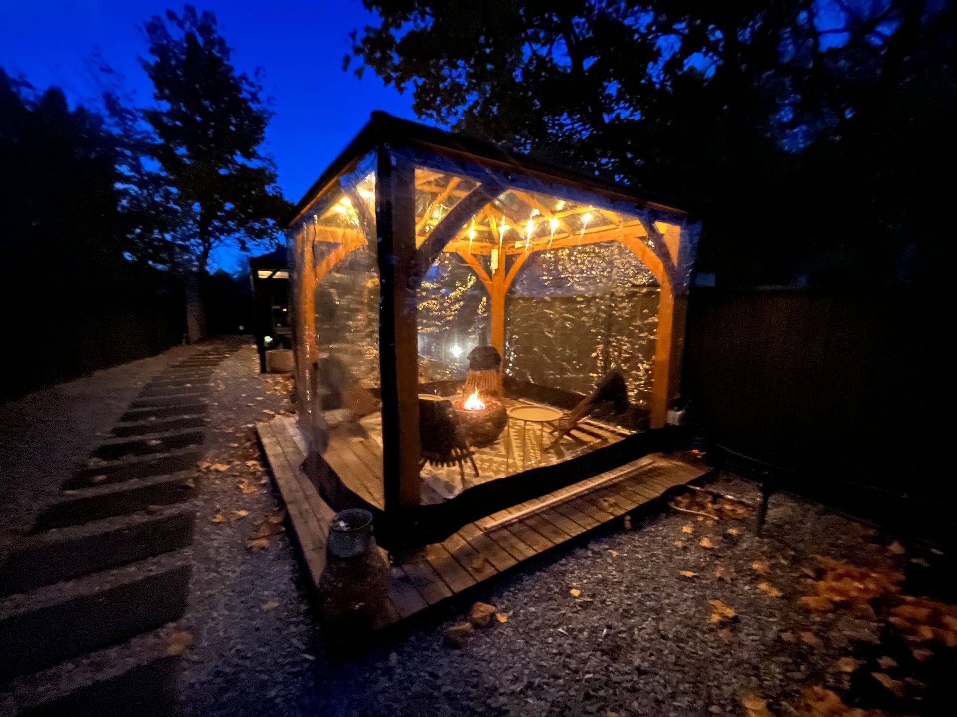 An outdoor wooden gazebo with clear walls, illuminated by warm string lights and a glowing fire pit at dusk.