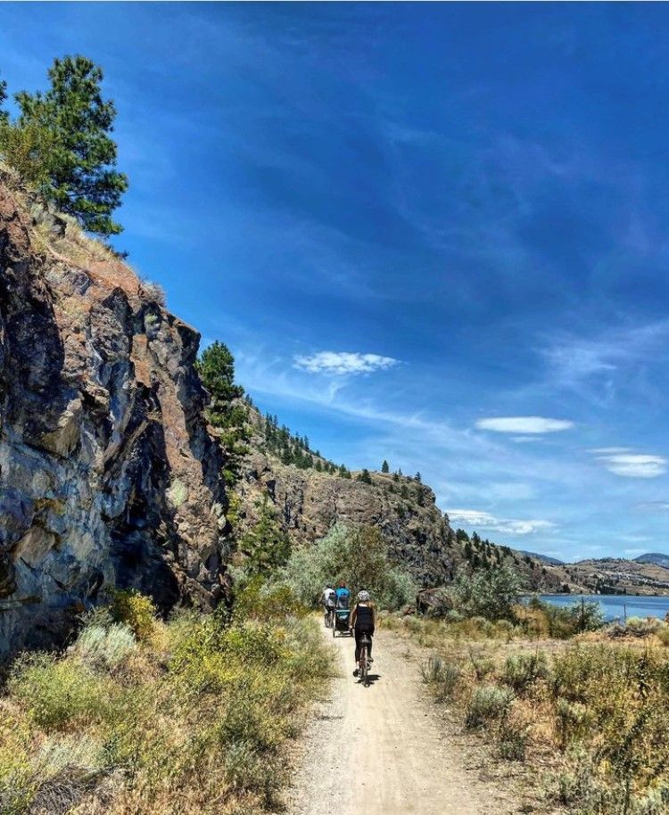 A person rides a bicycle along a dirt path beside a rocky cliff and a lake under a bright blue sky.
