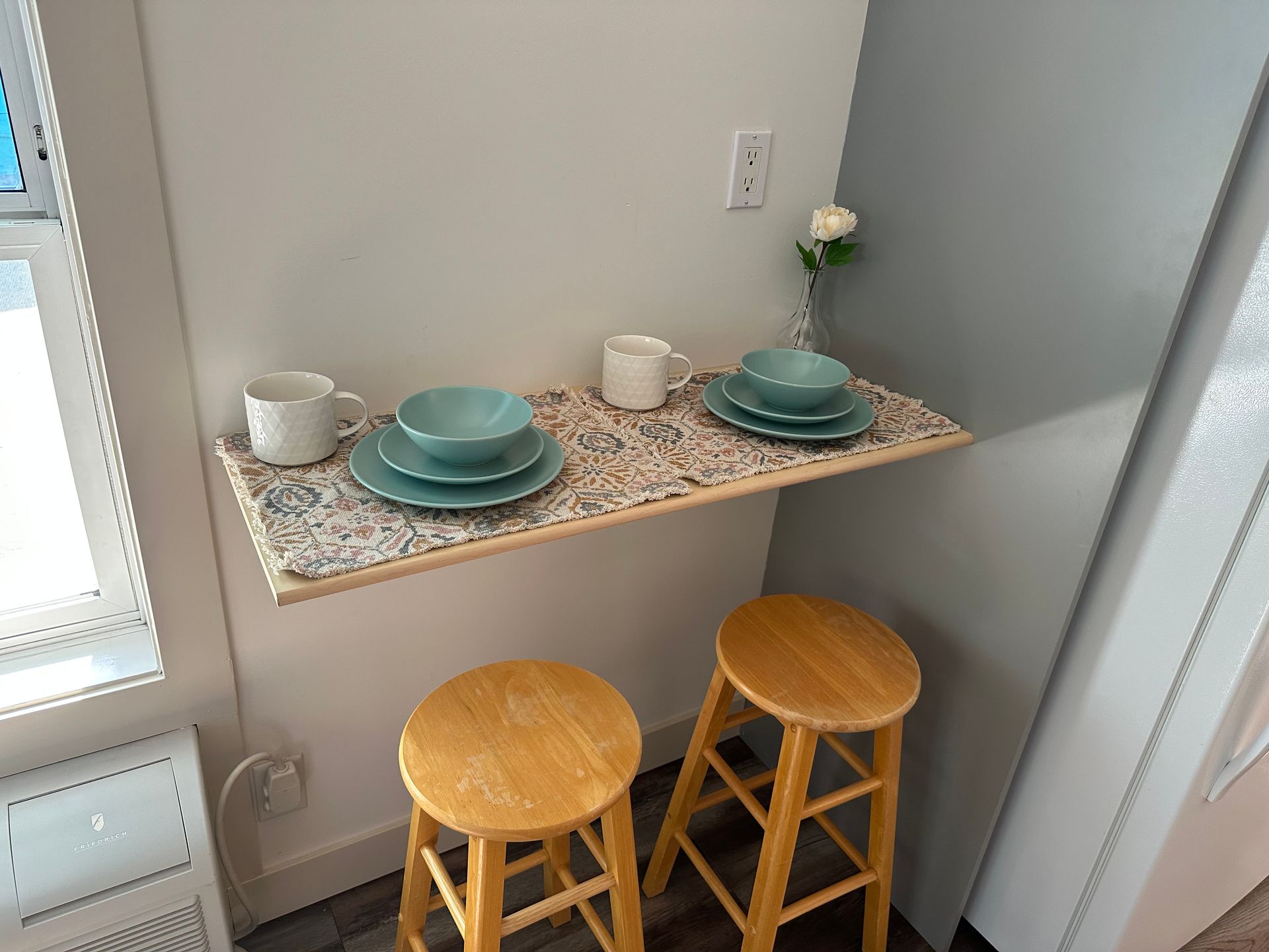 A small, wall-mounted wooden table set for two with blue dishes, mugs, and a single white rose between two wooden stools.