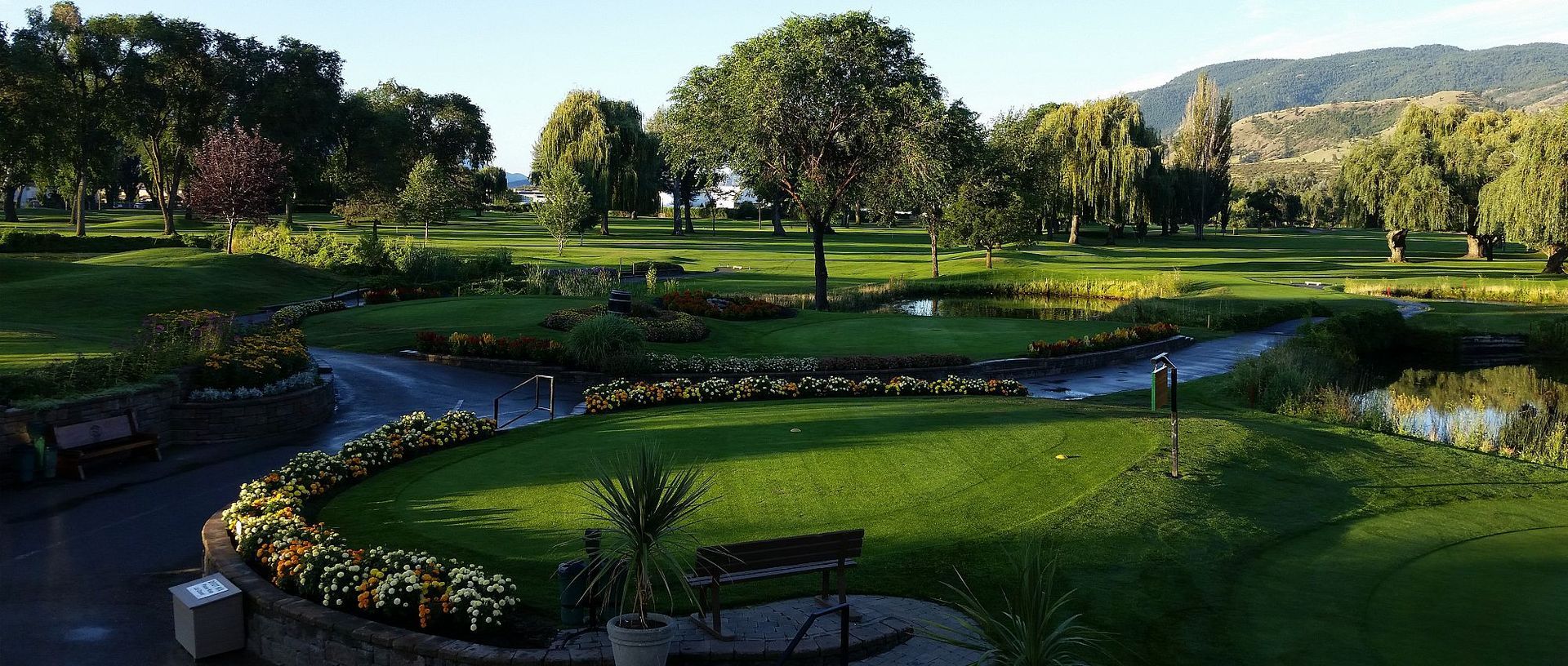 A lush green park with landscaped flower beds, winding pathways, a small pond, and benches under trees at sunset.