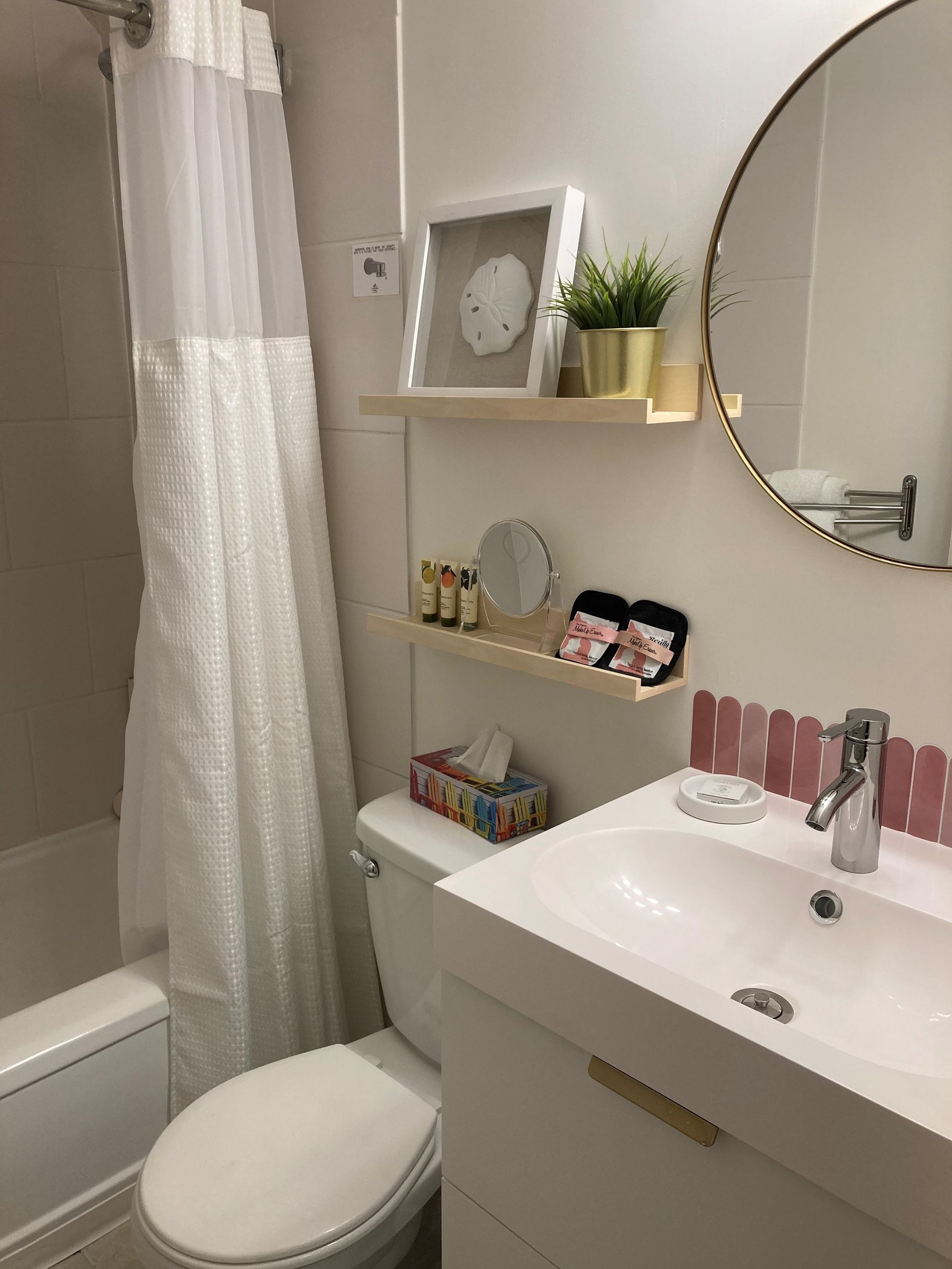 A white bathroom with a shower curtain, toilet, sink vanity with gold hardware, and two floating shelves with decor.