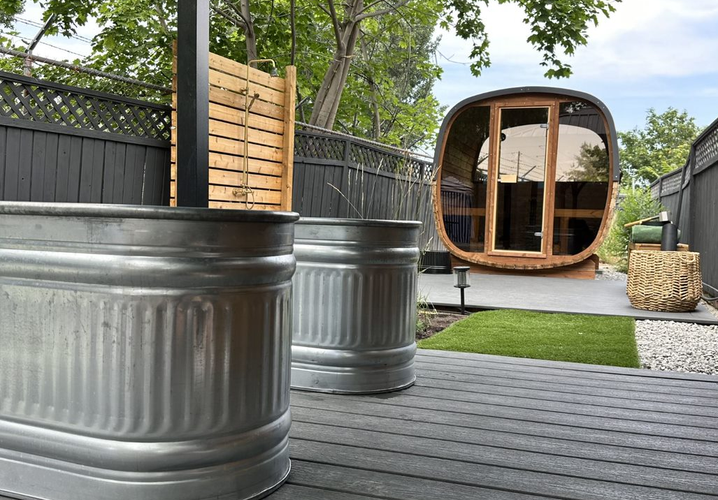 Two metal troughs sit on a deck near a wooden barrel sauna in a backyard with a fence and artificial grass.