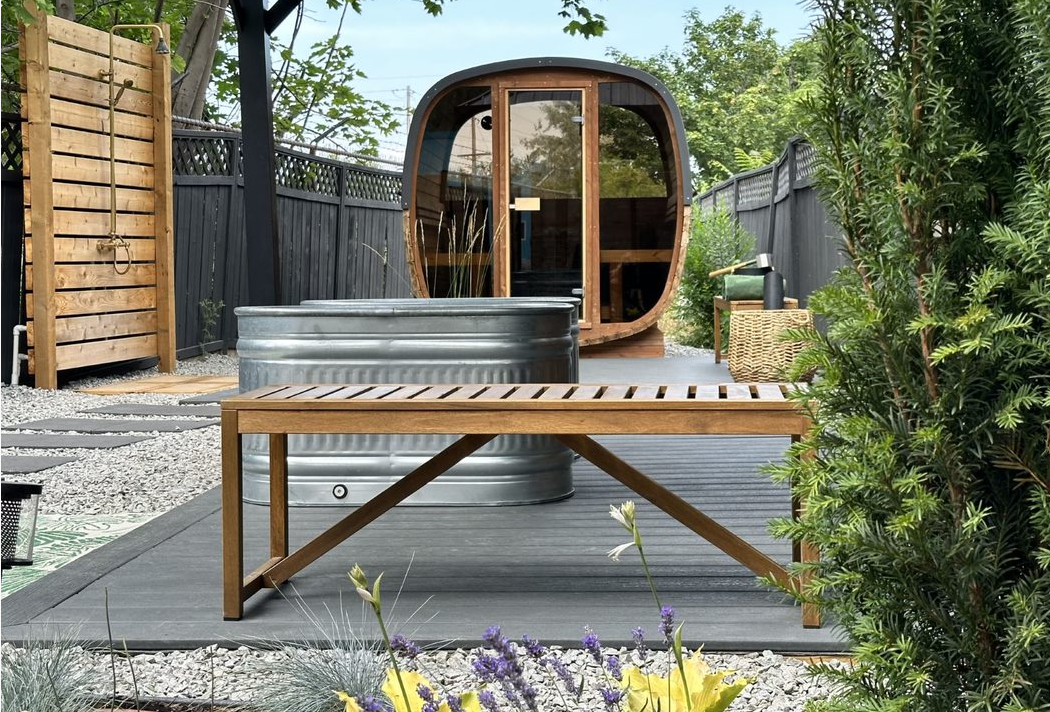 A wooden bench sits on a dark deck in front of a metal stock tank and a barrel sauna in a landscaped backyard.