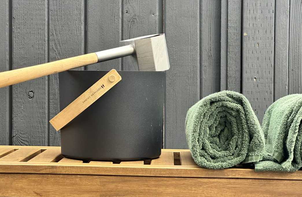 A black sauna bucket with a wooden handle, a metal ladle, and two rolled green towels on a wooden bench.