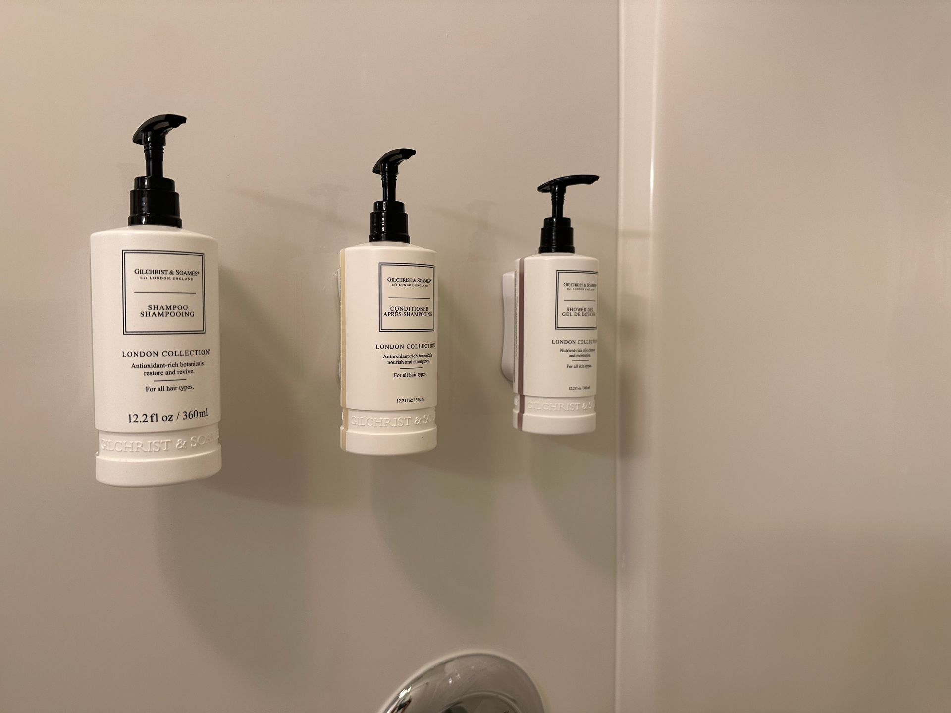 Three white pump bottles of hotel toiletries mounted on a beige shower wall.