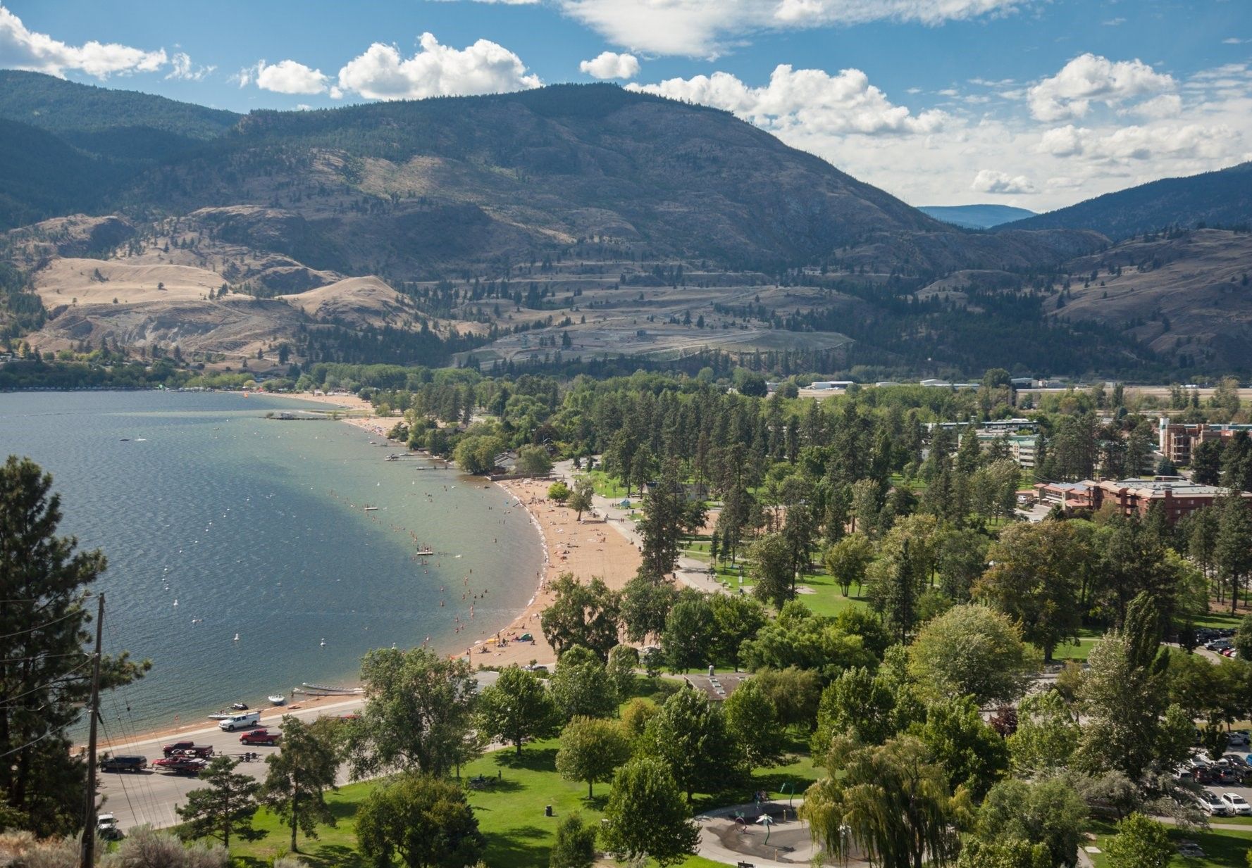 A wide-angle view of a sandy beach curving along a large lake, bordered by lush green trees and rugged hills in the back.
