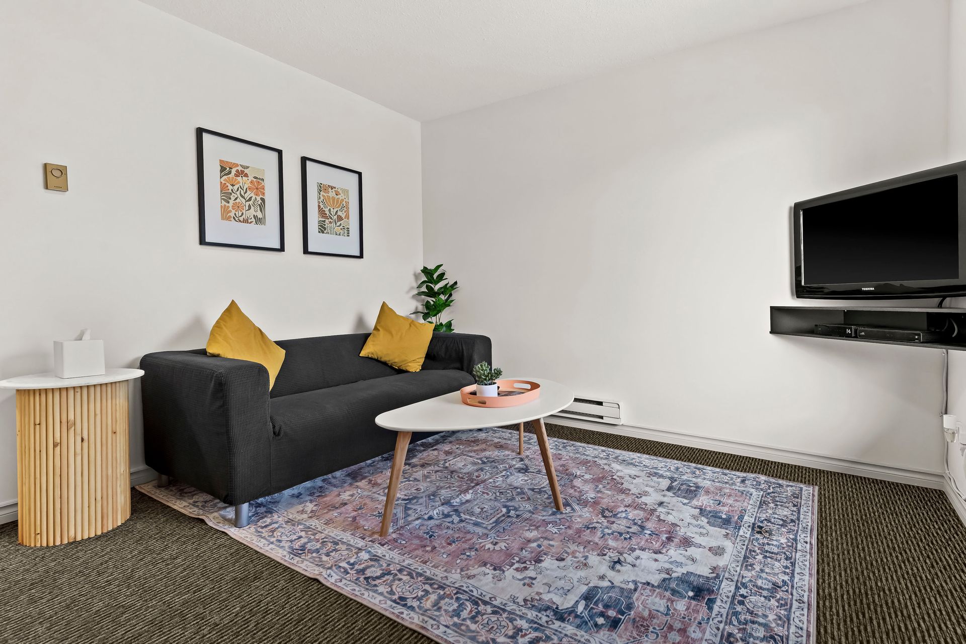 A living room with a dark sofa, two yellow pillows, a coffee table on a patterned rug, and a wall-mounted TV.