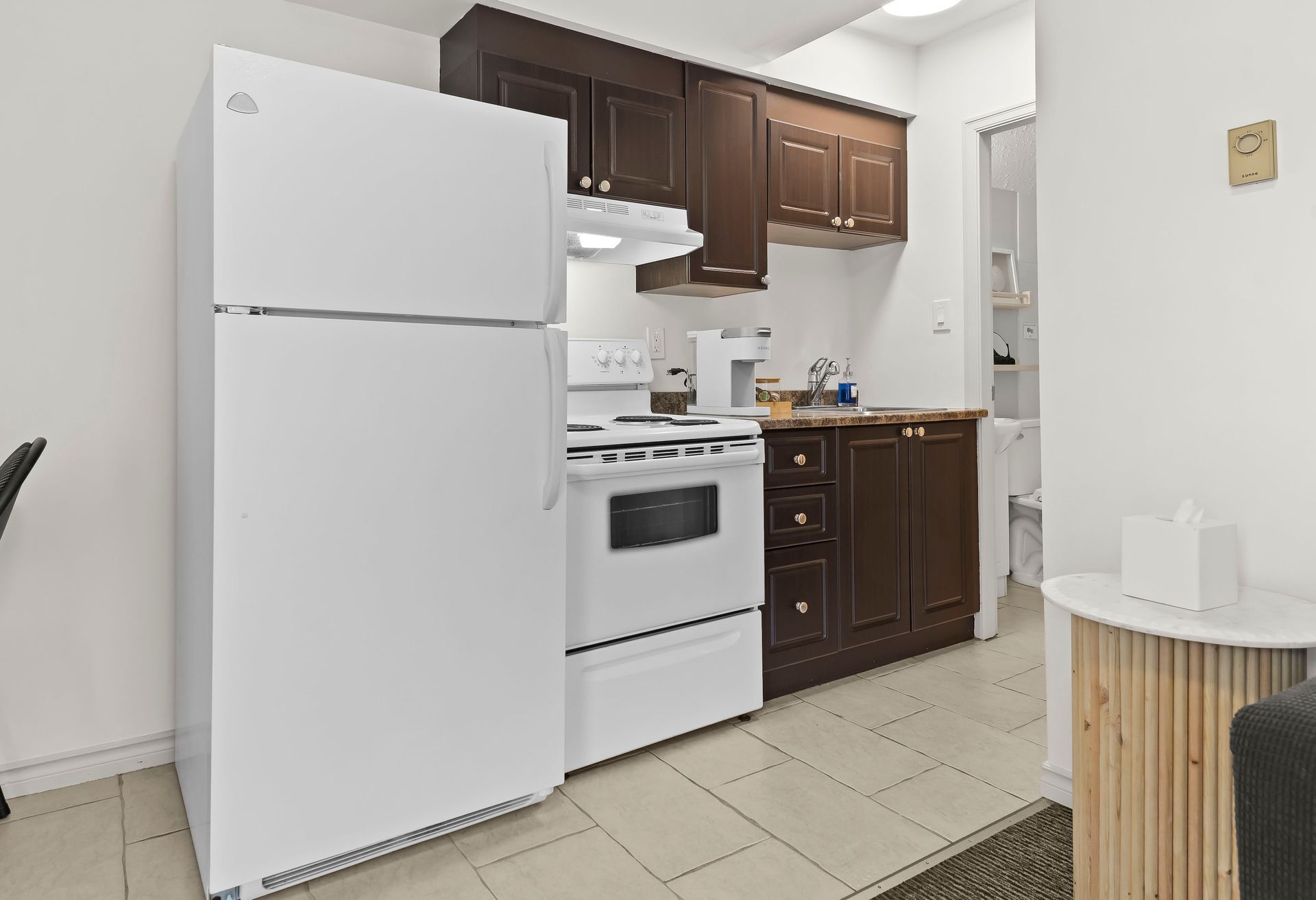 A small kitchen featuring a white refrigerator and stove, dark wood cabinets, and light tile flooring.