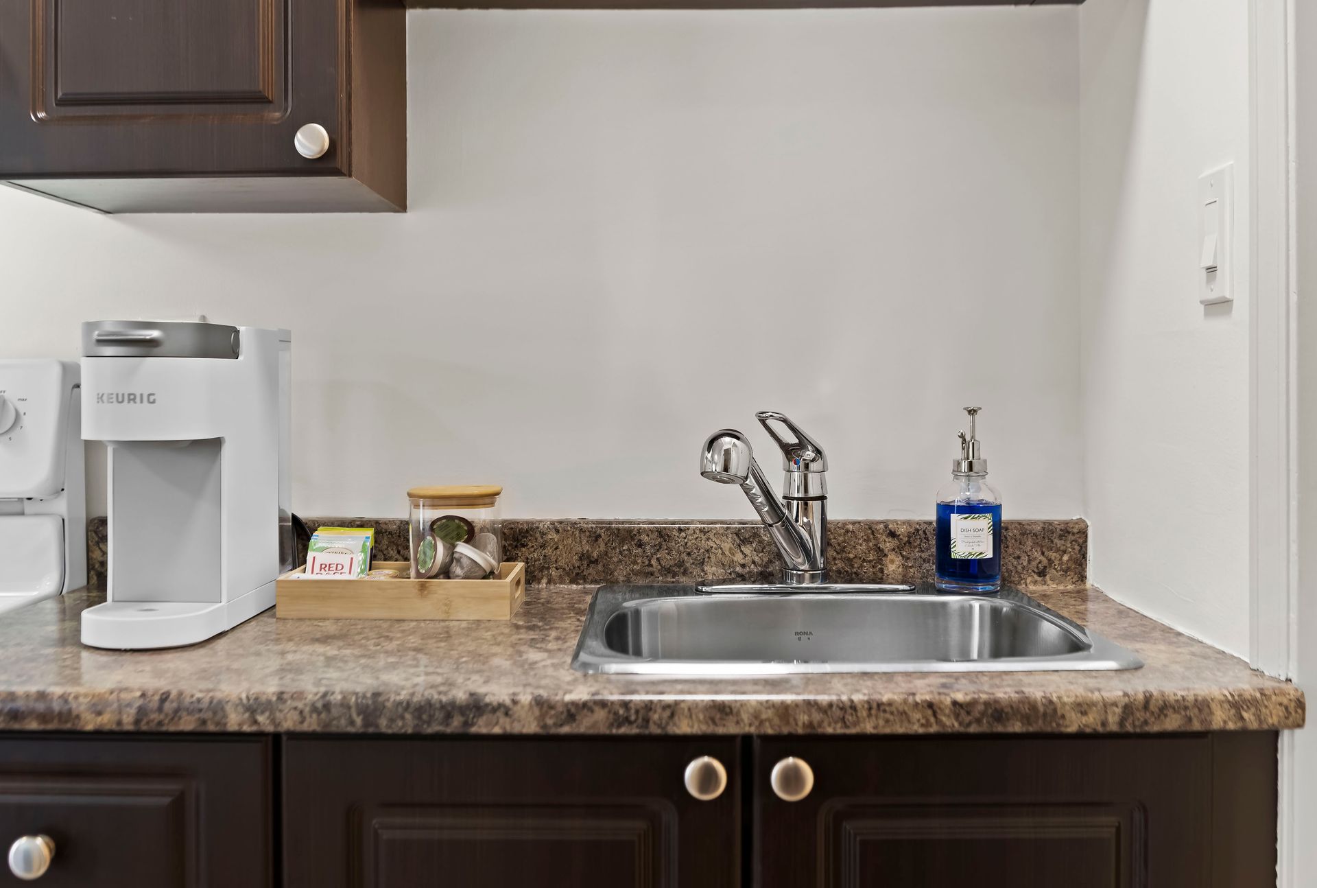 A white coffee maker, a wooden tray with supplies, and a chrome faucet set over a stainless sink on a brown countertop.