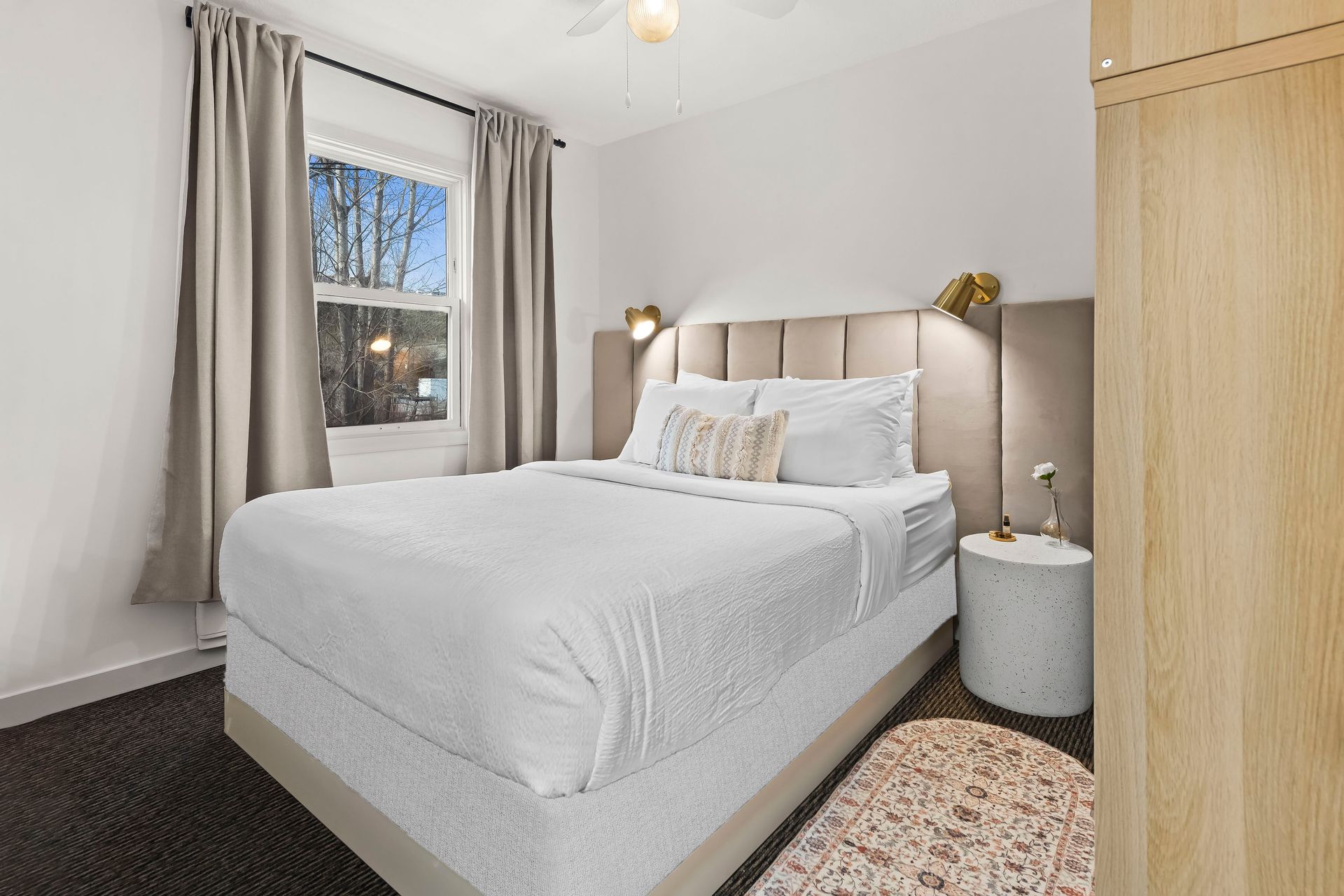 A bright bedroom with a queen bed featuring a beige tufted headboard, white bedding, gray curtains, and a stone nightstand.