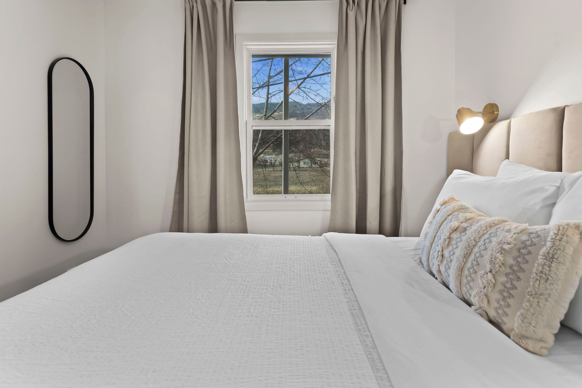 A clean, minimalist bedroom featuring a white bed, beige curtains, an oval black mirror, and a small wall-mounted light.