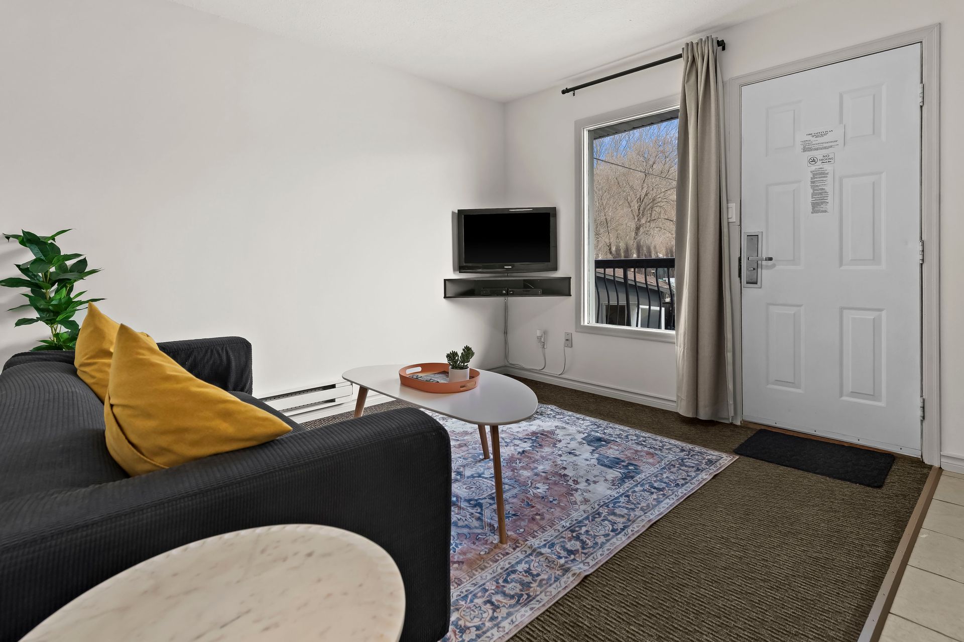 A cozy living room with a dark sofa, yellow cushions, a coffee table on a patterned rug, and a corner-mounted TV.