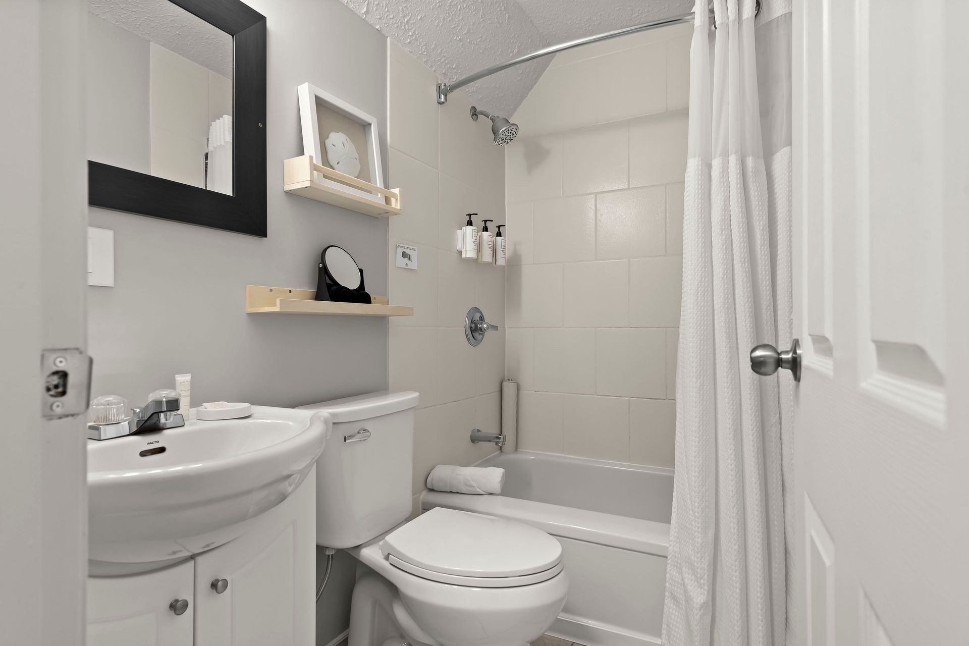 A small, bright bathroom with a white pedestal sink, a toilet, and a bathtub with a white curtain and wall-mounted bottles.