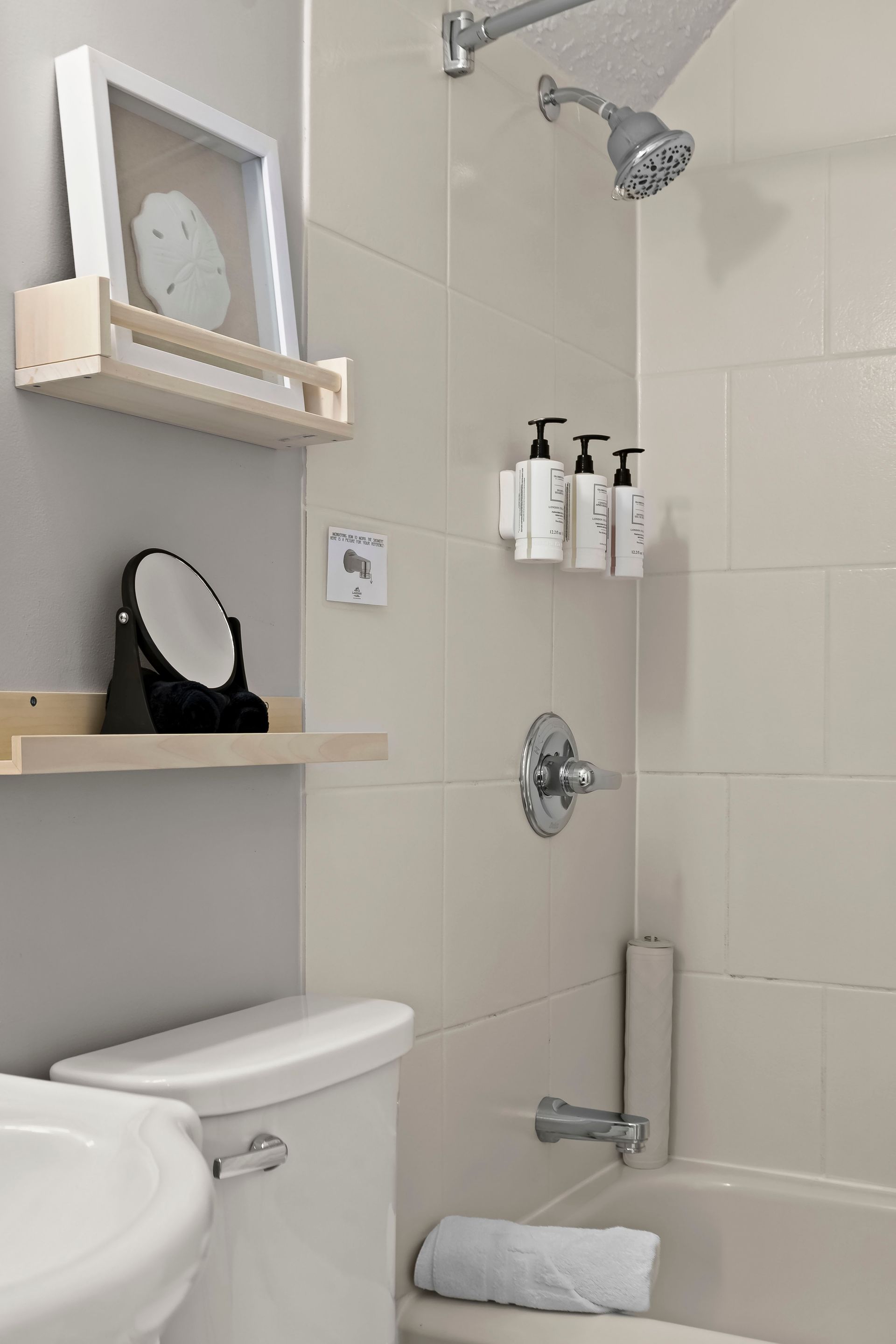 A clean, beige-tiled bathroom with a bathtub, shower, white toilet, and wooden shelves holding decor and a small mirror.