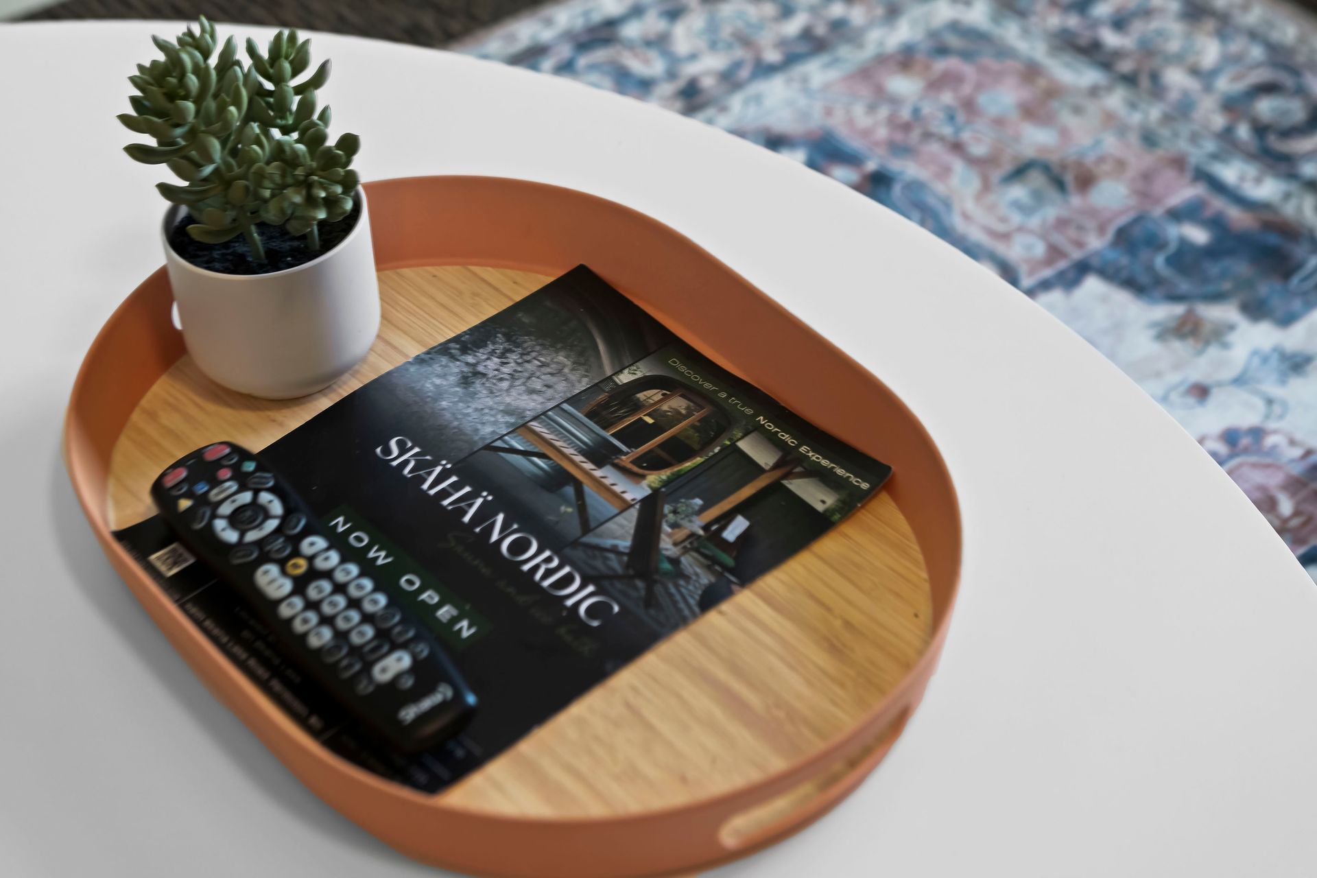 An oval wooden tray with a terracotta rim sits on a white table, holding a small plant, a remote, and a brochure.