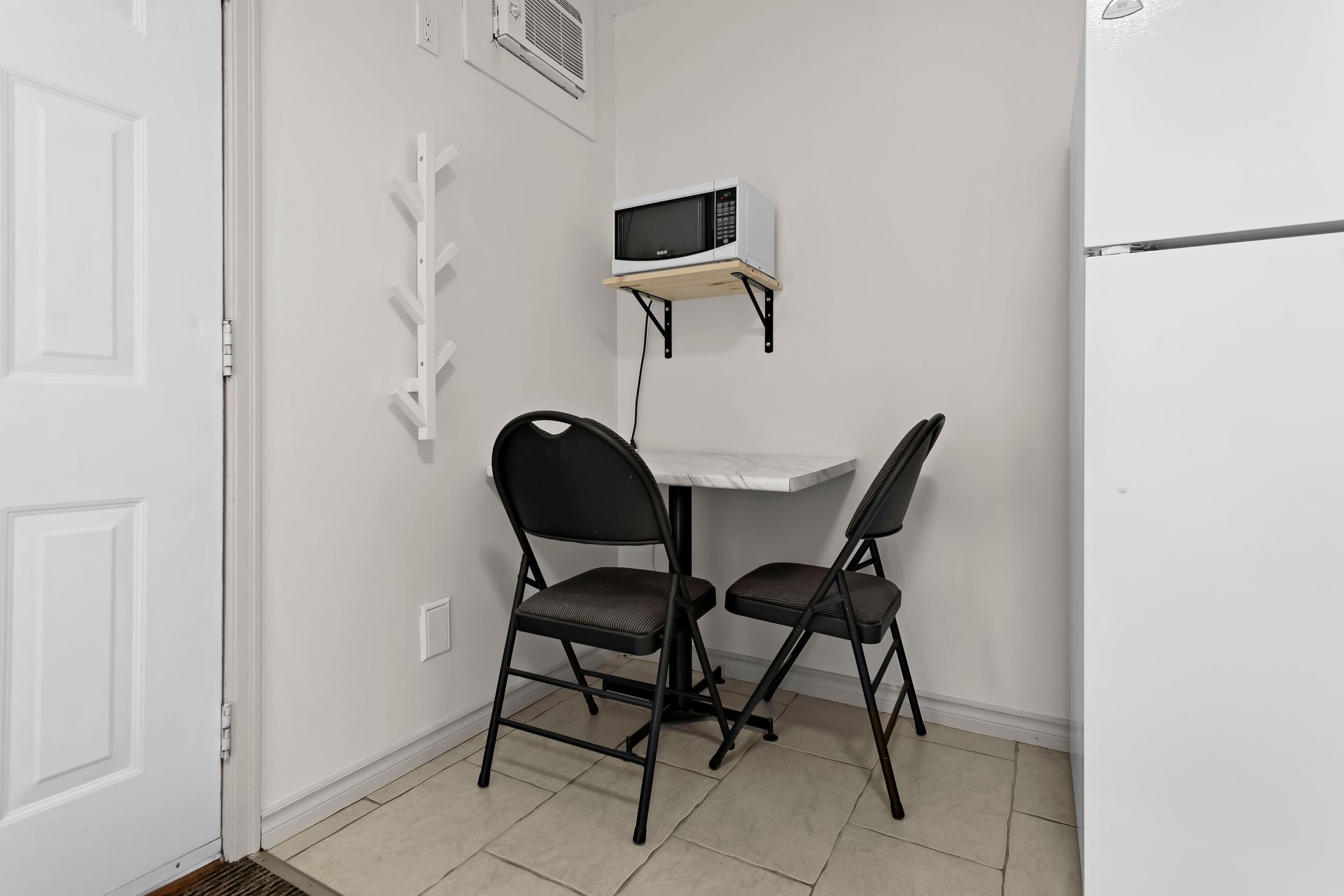A small dining area with two folding chairs at a small table, with a wall-mounted microwave above, on a tiled floor.