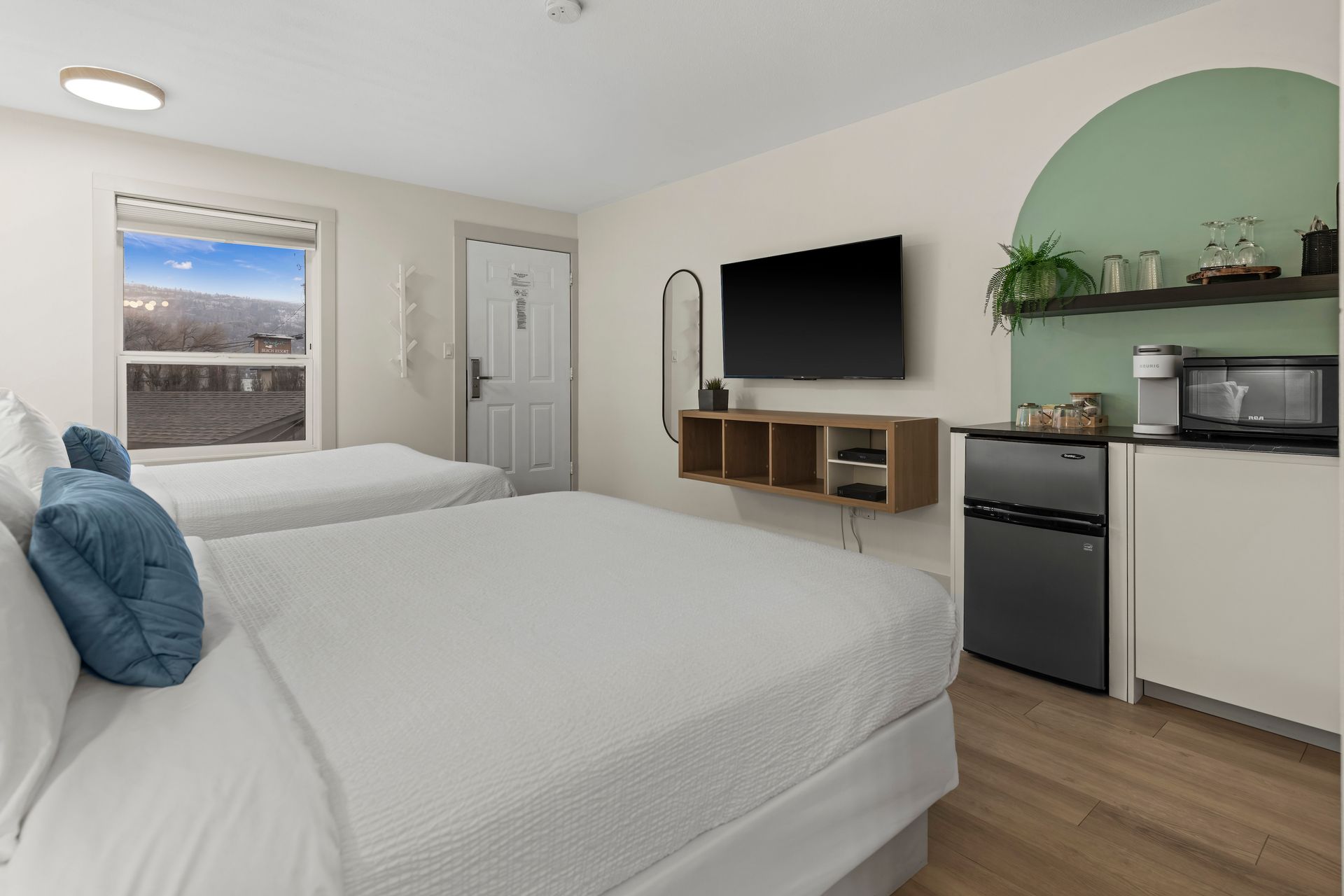 A hotel room with two white-bedded beds, a wall-mounted television, and a kitchenette area featuring a green arched wall.