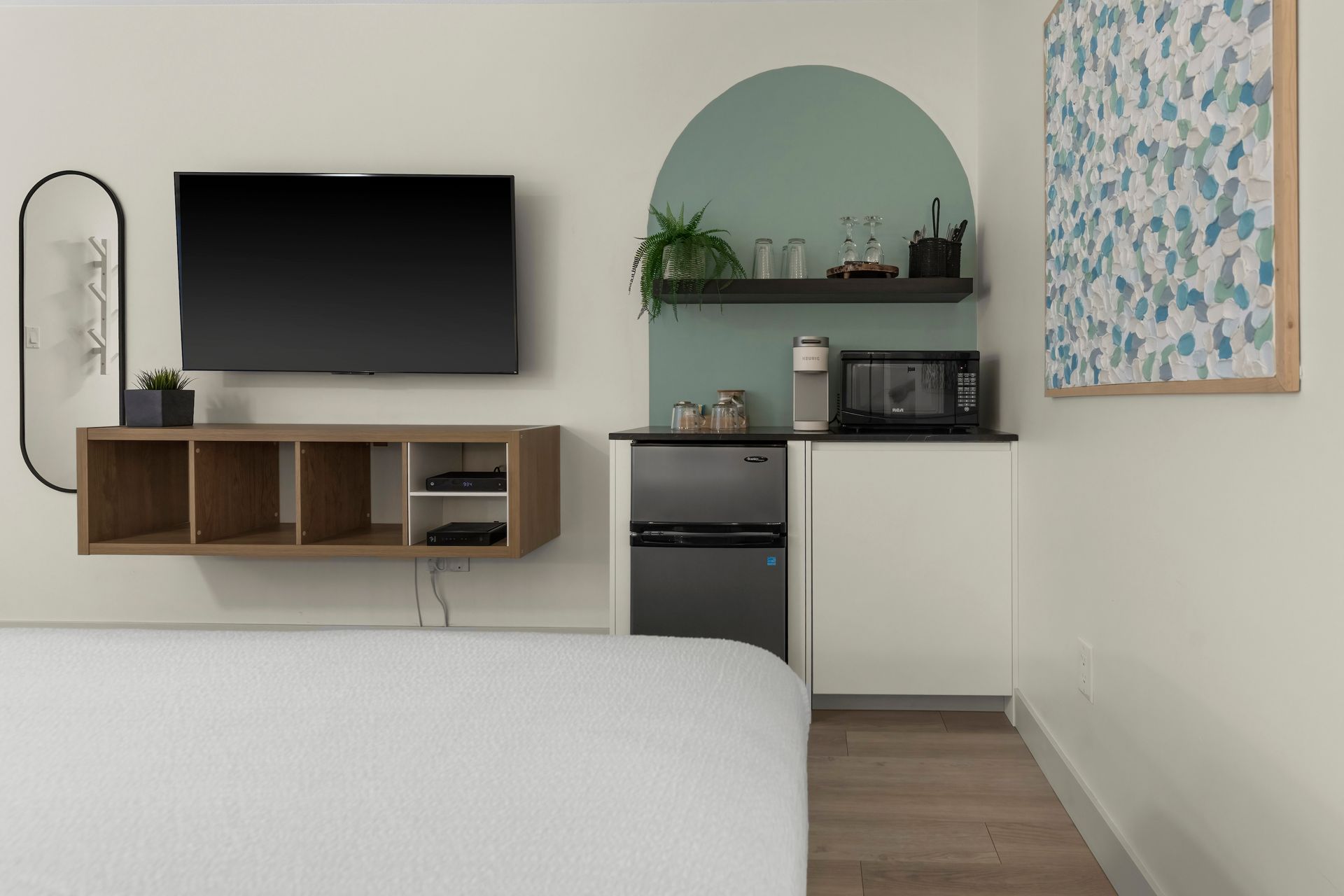 A modern bedroom area featuring a wall-mounted TV above shelving, a minibar, microwave, and green arched accent wall.