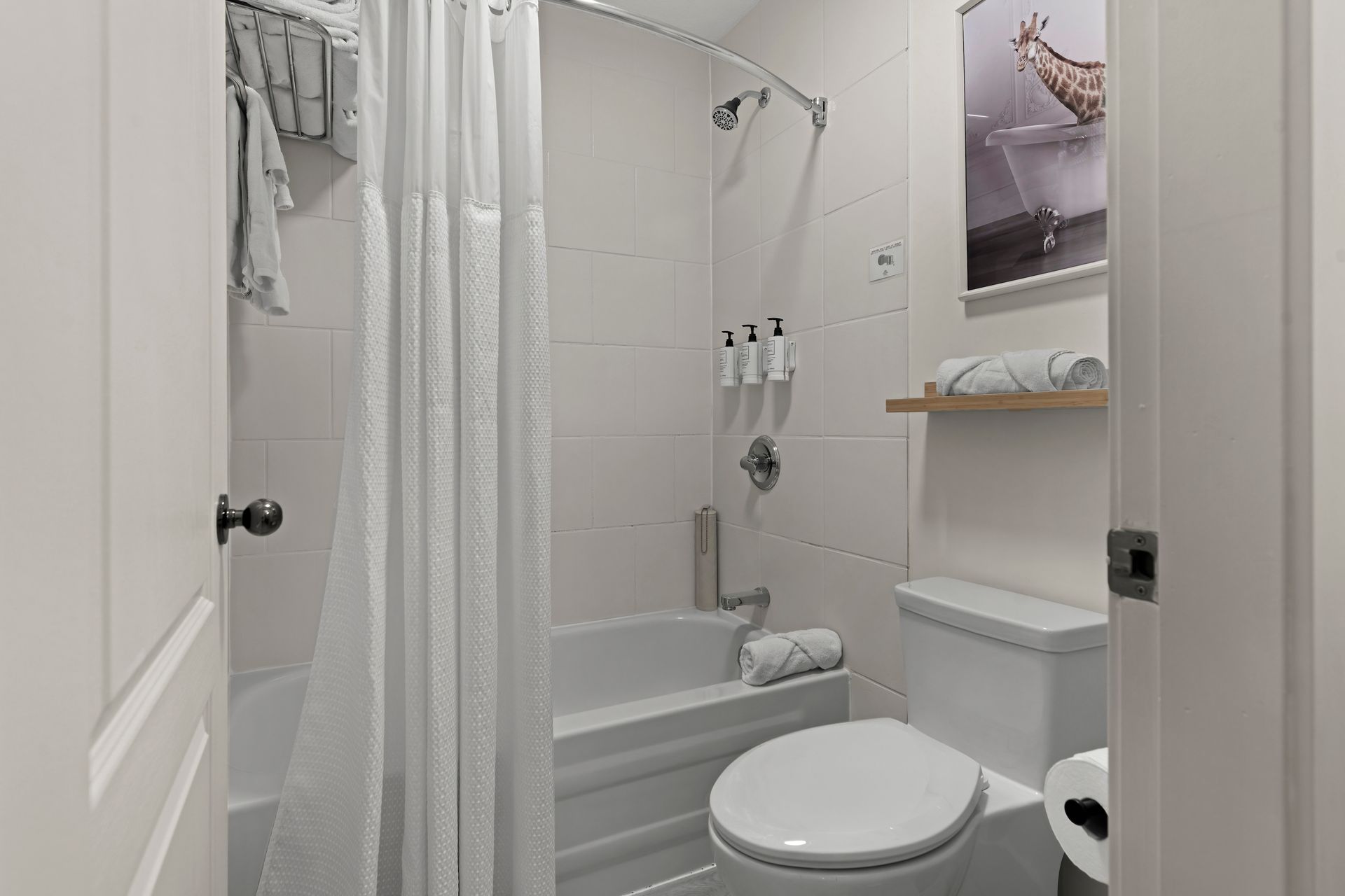 A bright, neutral-toned hotel bathroom featuring a shower-tub combo with a white curtain, a toilet, and wall decor.