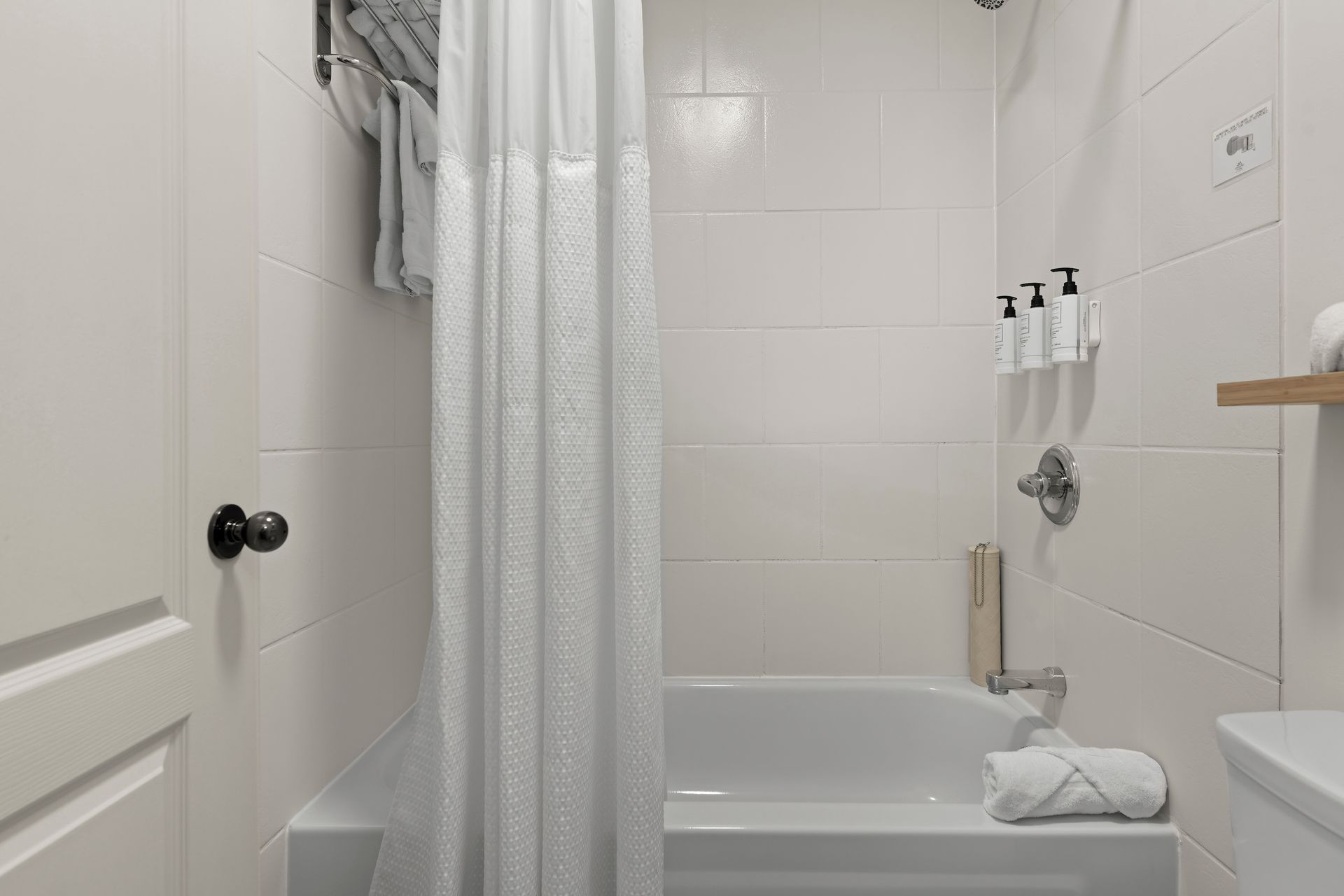 A white tiled bathroom featuring a bathtub, shower curtain, mounted pump bottles, and folded white towels.