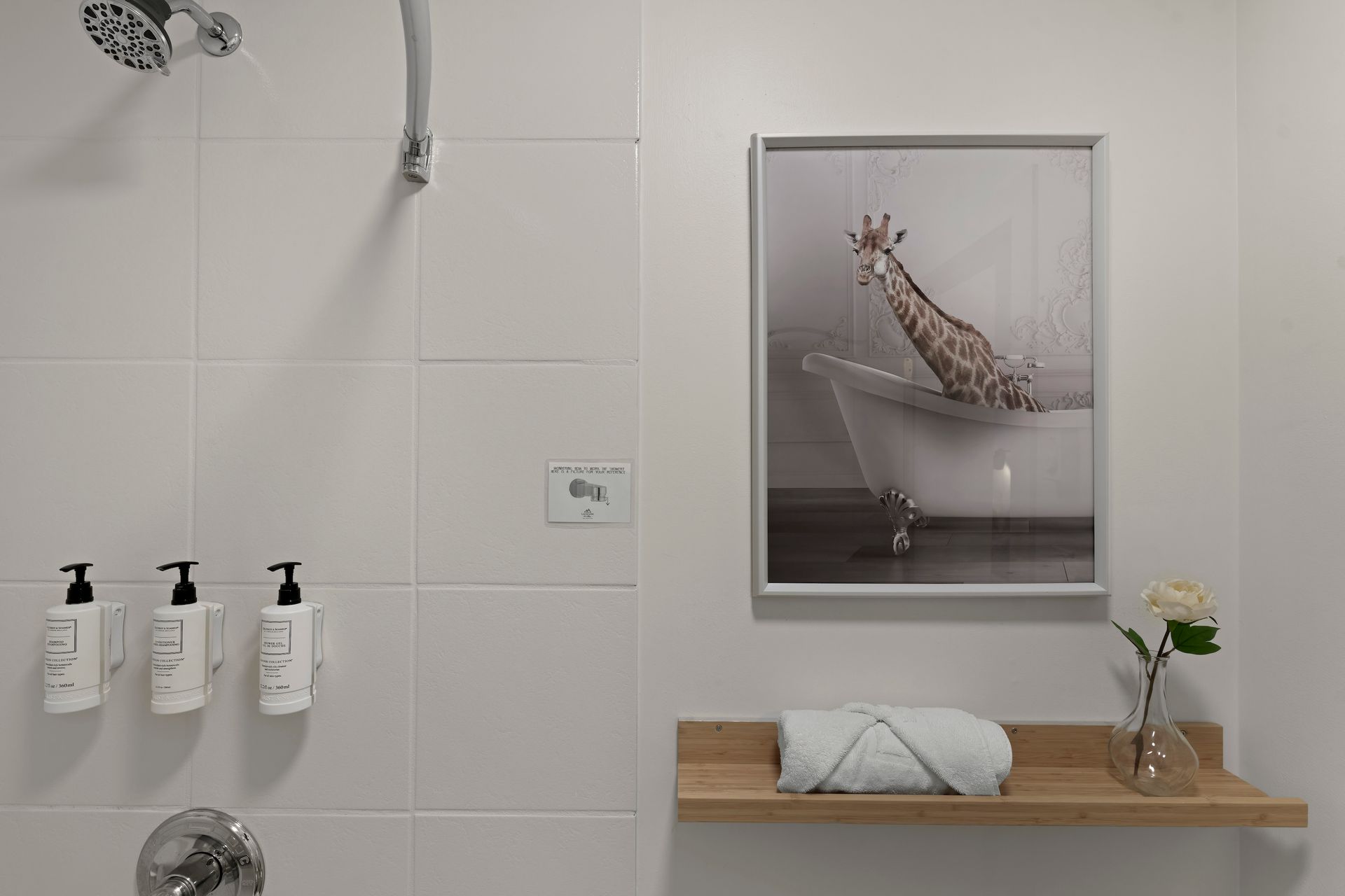 A white-tiled shower area with wall-mounted soap dispensers, a framed print of a giraffe in a tub, and a towel on a shelf.