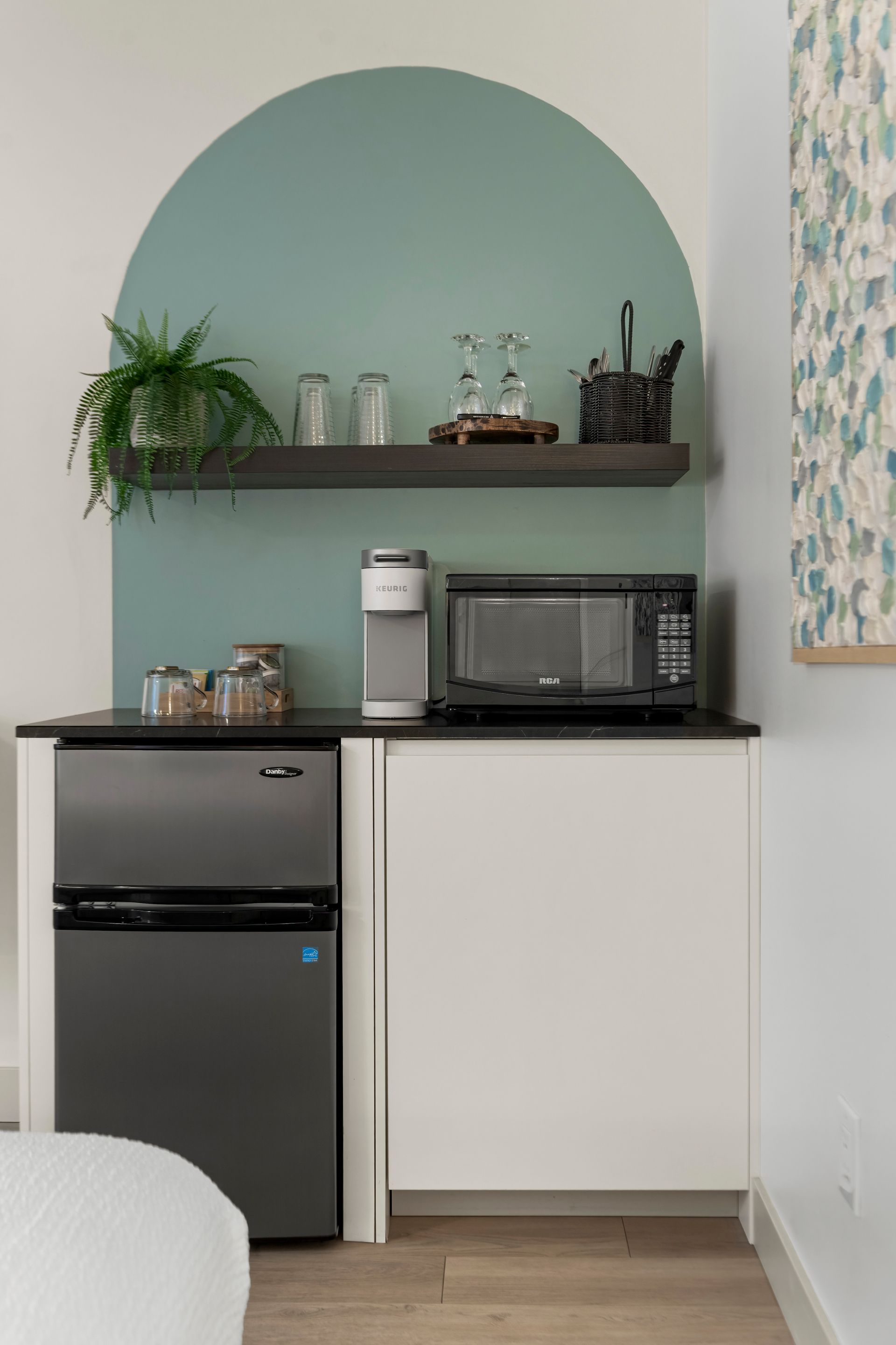 A white cabinet with a mini fridge, microwave, and coffee maker, featuring a sage green painted arch and shelf on the wall.