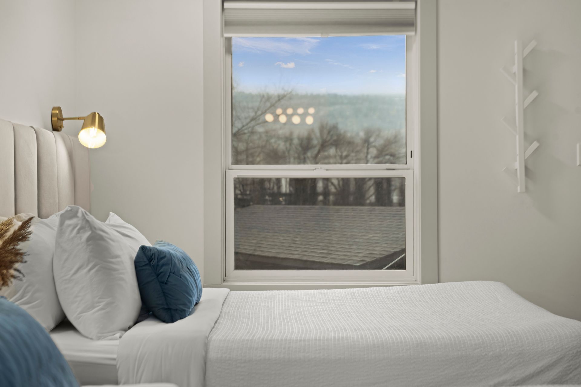 A bedroom with a bed featuring white bedding, blue and white pillows, a brass wall lamp, and a view of trees via a window.